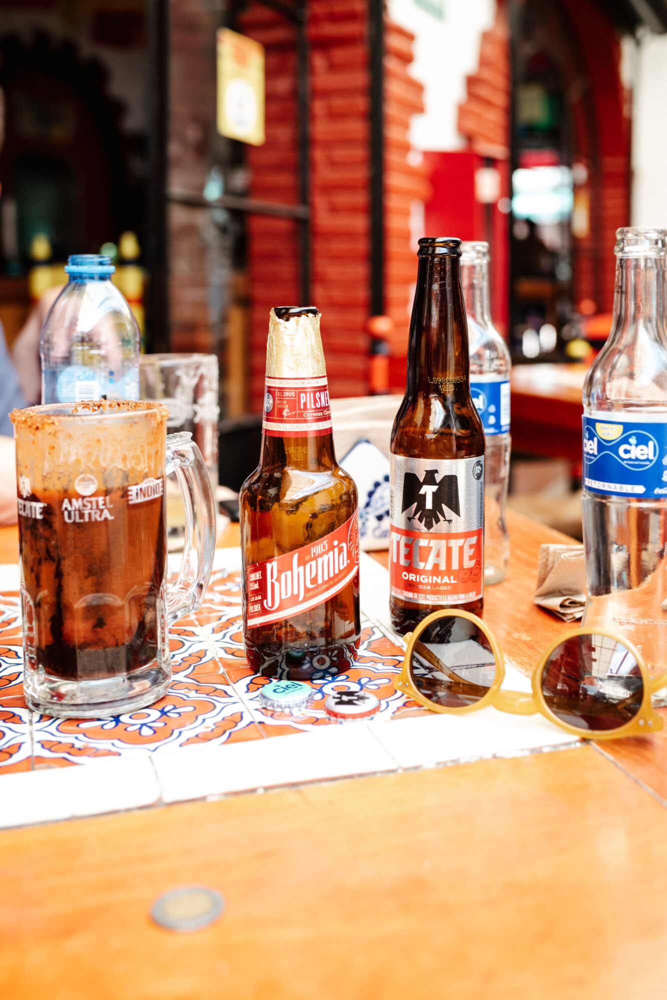 Seattle's best commercial food and hospitality photographer A table captured by Seattles best commercial food lifestyle photographer displays Bohemia and Tecate beer, Ciel water, a chili-rimmed mug, and sunglasses on a vibrant tiled surface at an outdoor restaurant. captured by seattle's best food and lifestyle photographer Brooke Fitts
