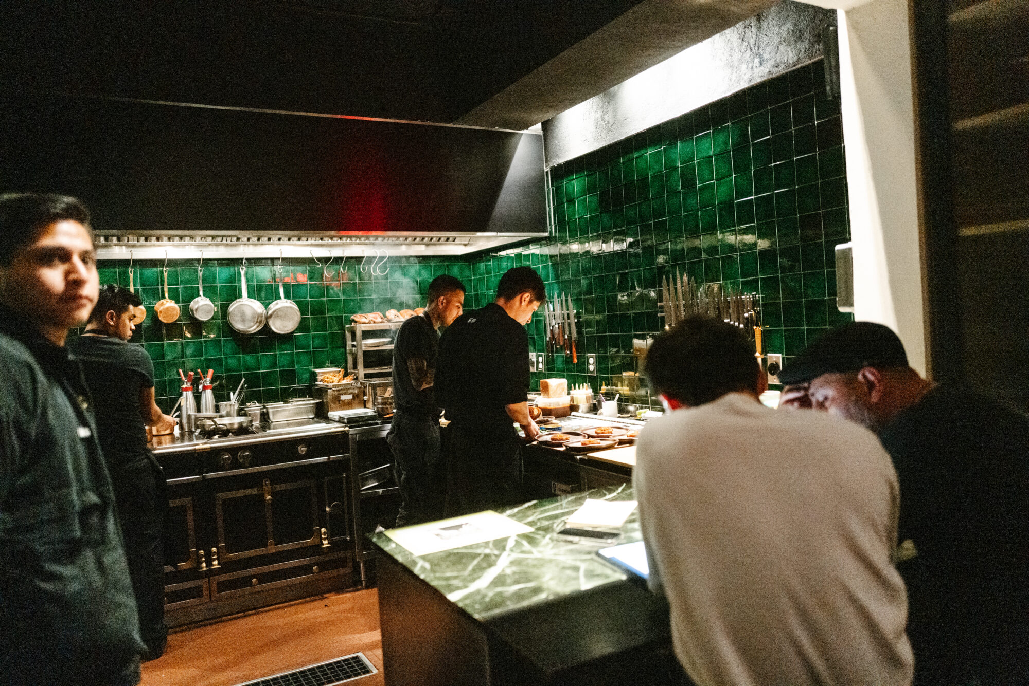 Seattle's best commercial food and hospitality photographer Several people work in a restaurant kitchen with green tiled walls, preparing food at a counter and stove, while two others look at paperwork. Captured by Seattles best commercial food lifestyle photographer, pots and utensils hang on the wall. captured by seattle's best food and lifestyle photographer Brooke Fitts