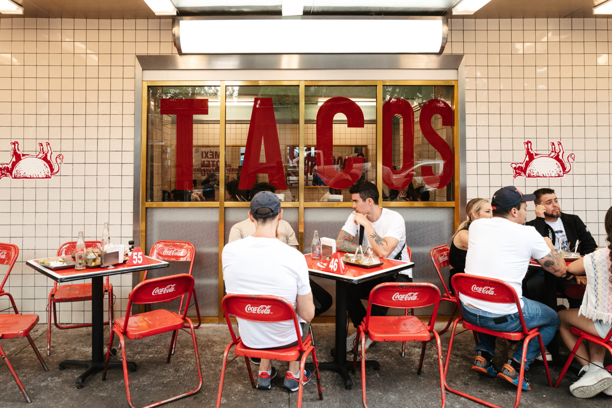 Seattle's best commercial food and hospitality photographer People sit at red Coca-Cola branded chairs outside a tiled restaurant with large red letters spelling TACOS on the window, captured by Seattles best commercial food lifestyle photographer. Diners are eating, talking, and enjoying the casual vibe. captured by seattle's best food and lifestyle photographer Brooke Fitts