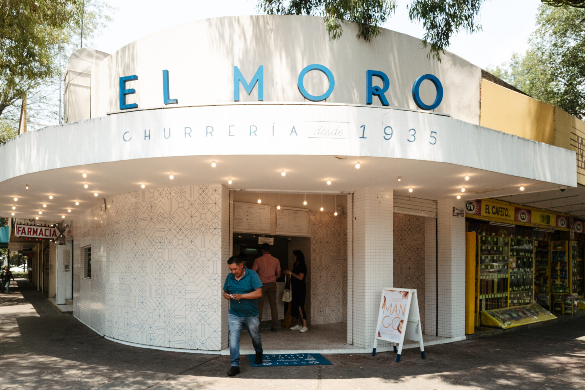 Seattle's best commercial food and hospitality photographer A man exits the white, round-cornered El Moro churrería, established in 1935, with blue signage on the facade. Captured by Seattles best commercial food lifestyle photographer, two women stand at the entrance near a pharmacy and a yellow storefront. captured by seattle's best food and lifestyle photographer Brooke Fitts