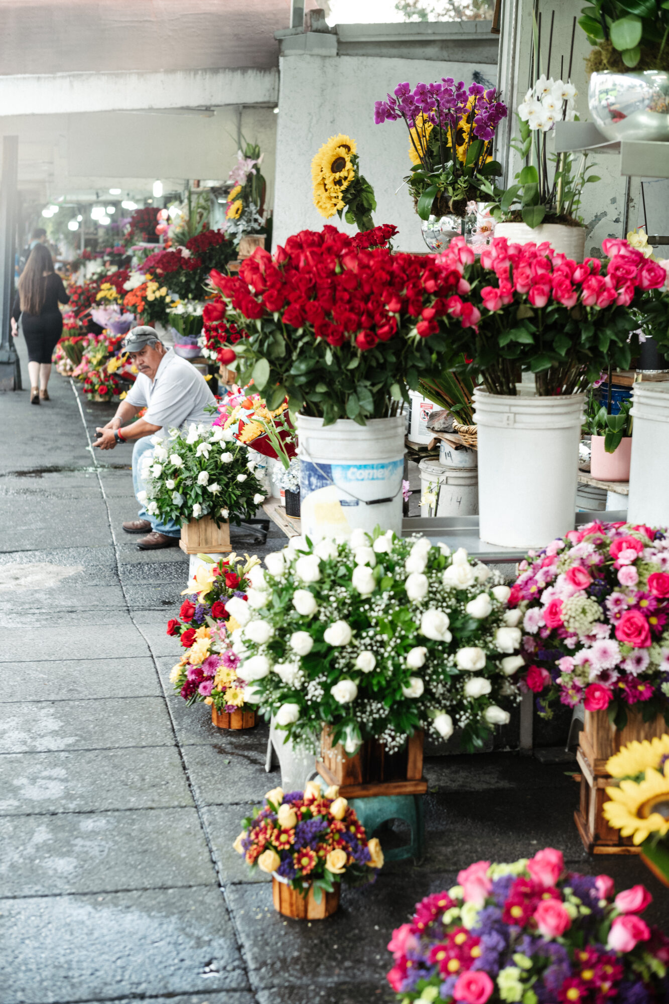 Seattle's best commercial food and hospitality photographer A sidewalk flower market displays vibrant bouquets in buckets and crates—captured by Seattles best commercial food lifestyle photographer. A man sits nearby, while a woman walks in the background among red roses, sunflowers, and more. captured by seattle's best food and lifestyle photographer Brooke Fitts