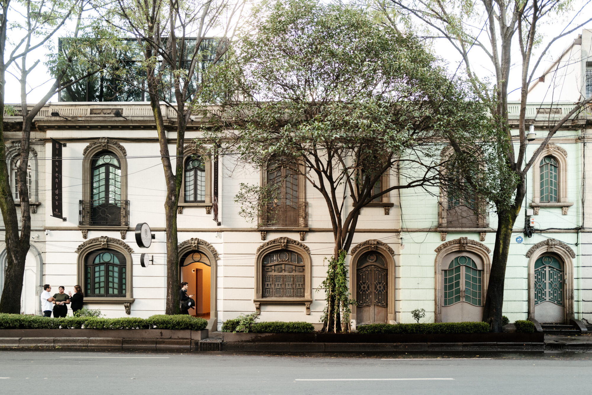 Seattle's best commercial food and hospitality photographer A row of old, ornate buildings with arched windows and doors is partially obscured by leafy trees. Four people sit on a bench to the left, while a person stands near an open orange door—captured by Seattles best commercial food lifestyle photographer. captured by seattle's best food and lifestyle photographer Brooke Fitts