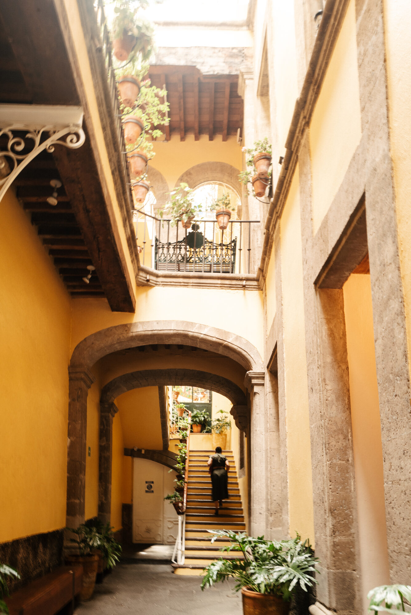 Seattle's best commercial food and hospitality photographer A bright, yellow-walled courtyard with stone arches, stairs leading up to a balcony lined with potted plants, and a person walking up the stairs—captured by Seattles best commercial food lifestyle photographer. Sunlight filters in, creating a warm and inviting atmosphere. captured by seattle's best food and lifestyle photographer Brooke Fitts