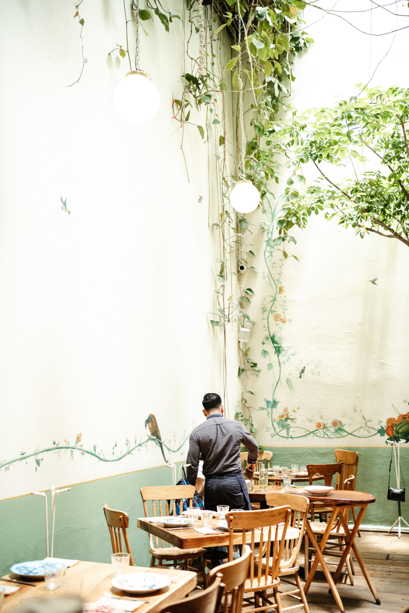 Seattle's best commercial food and hospitality photographer A waiter in a dark shirt cleans tables in a bright, airy café with wooden chairs, high ceilings, and green plants cascading down muraled walls—captured by Seattles best commercial food lifestyle photographer. captured by seattle's best food and lifestyle photographer Brooke Fitts