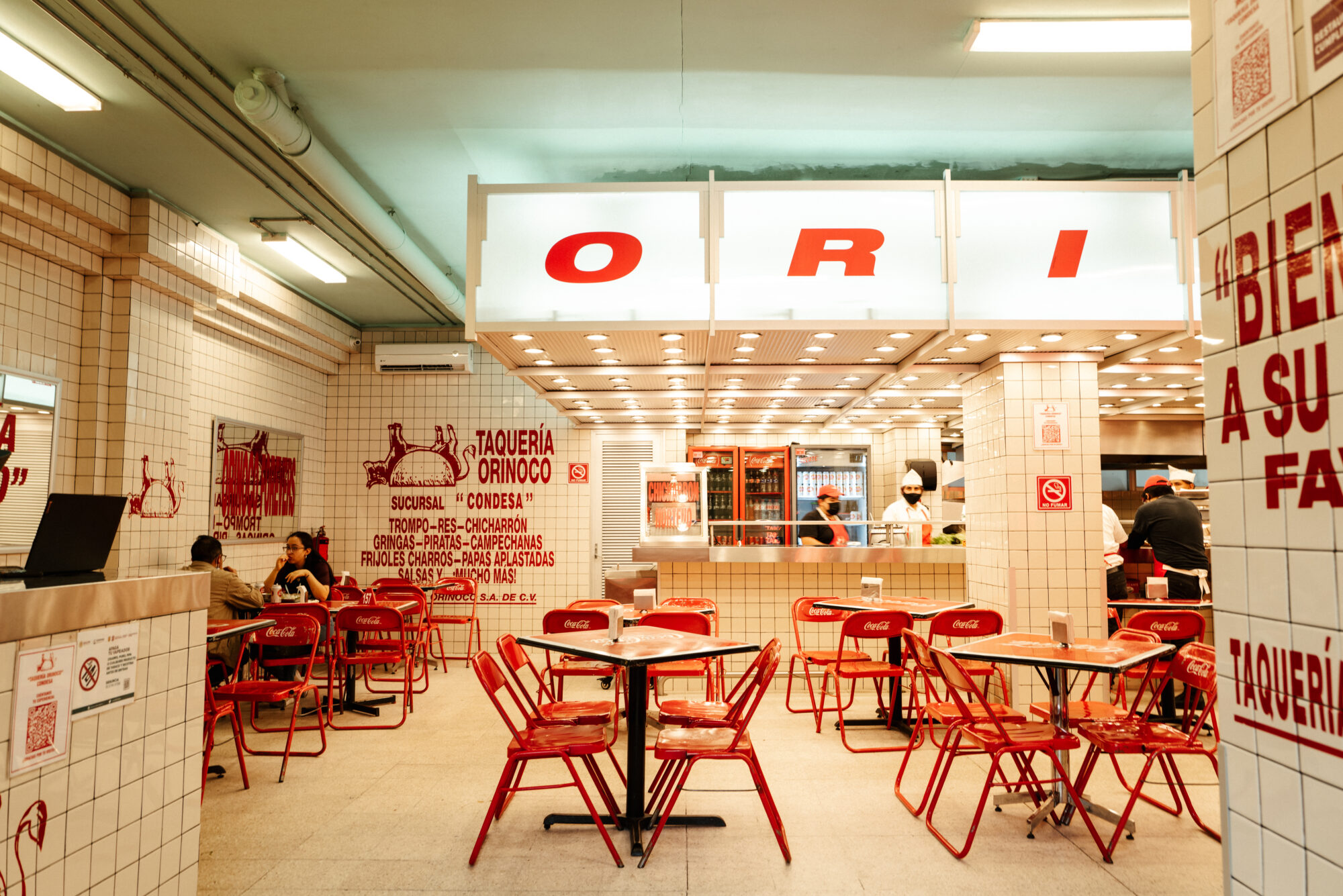 Seattle's best commercial food and hospitality photographer A taqueria with white tiled walls, red Coca-Cola chairs and tables, and bright overhead lights—captured perfectly by Seattles best commercial food lifestyle photographer. A few people are seated while a staff member works behind the counter amid vibrant red signage. captured by seattle's best food and lifestyle photographer Brooke Fitts