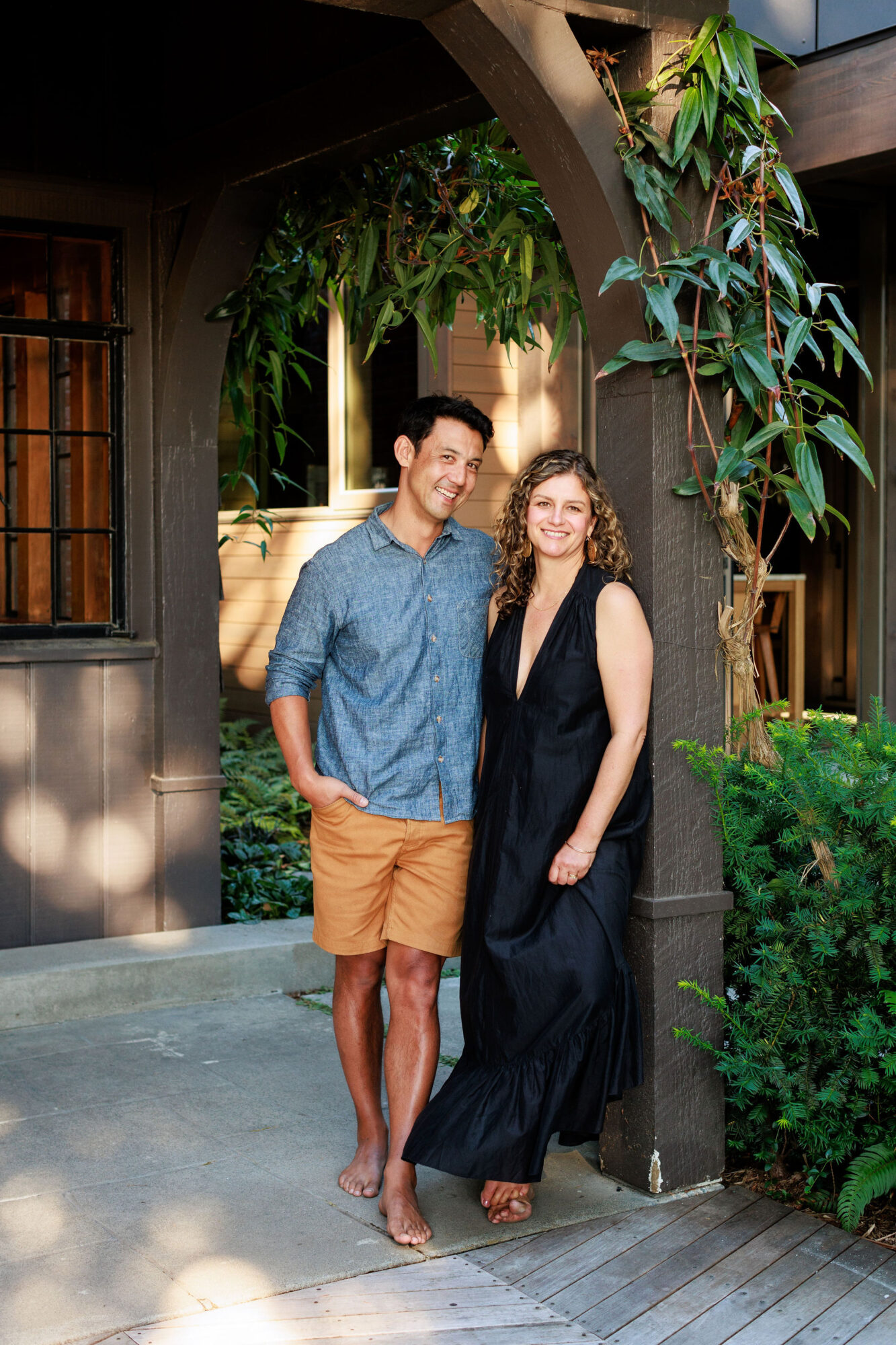 Seattle's best commercial food and hospitality photographer A smiling couple stands under a wooden arch covered with green vines. Captured by Seattles best commercial food lifestyle photographer, the man wears a blue shirt and tan shorts, hands in pockets. The woman, in a long black dress, leans against the arch. captured by seattle's best food and lifestyle photographer Brooke Fitts