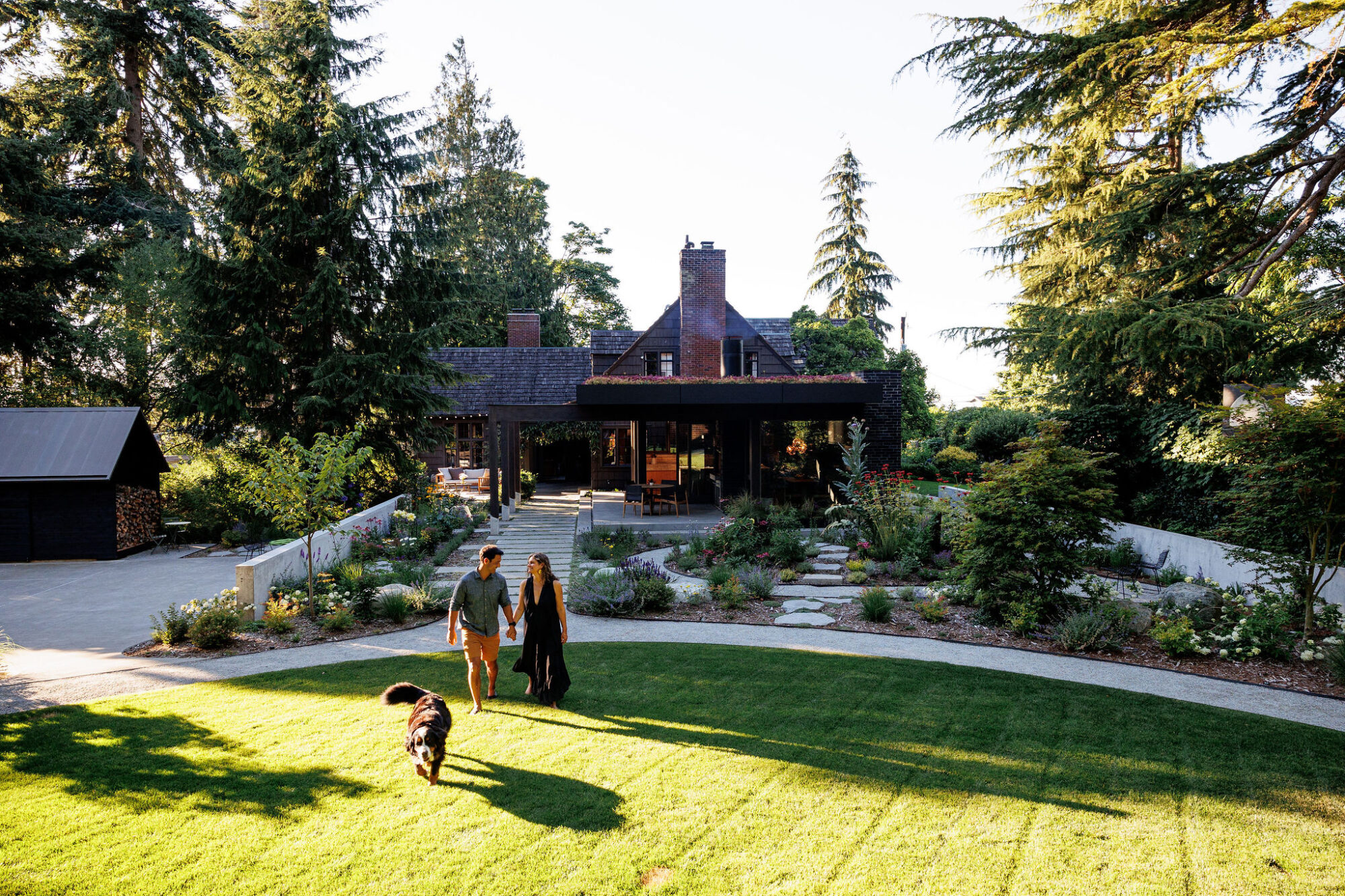 Seattle's best commercial food and hospitality photographer A couple walks with a black dog across a sunlit lawn toward a large, rustic house surrounded by tall trees and gardens—captured by Seattles best commercial food lifestyle photographer on a clear day. captured by seattle's best food and lifestyle photographer Brooke Fitts