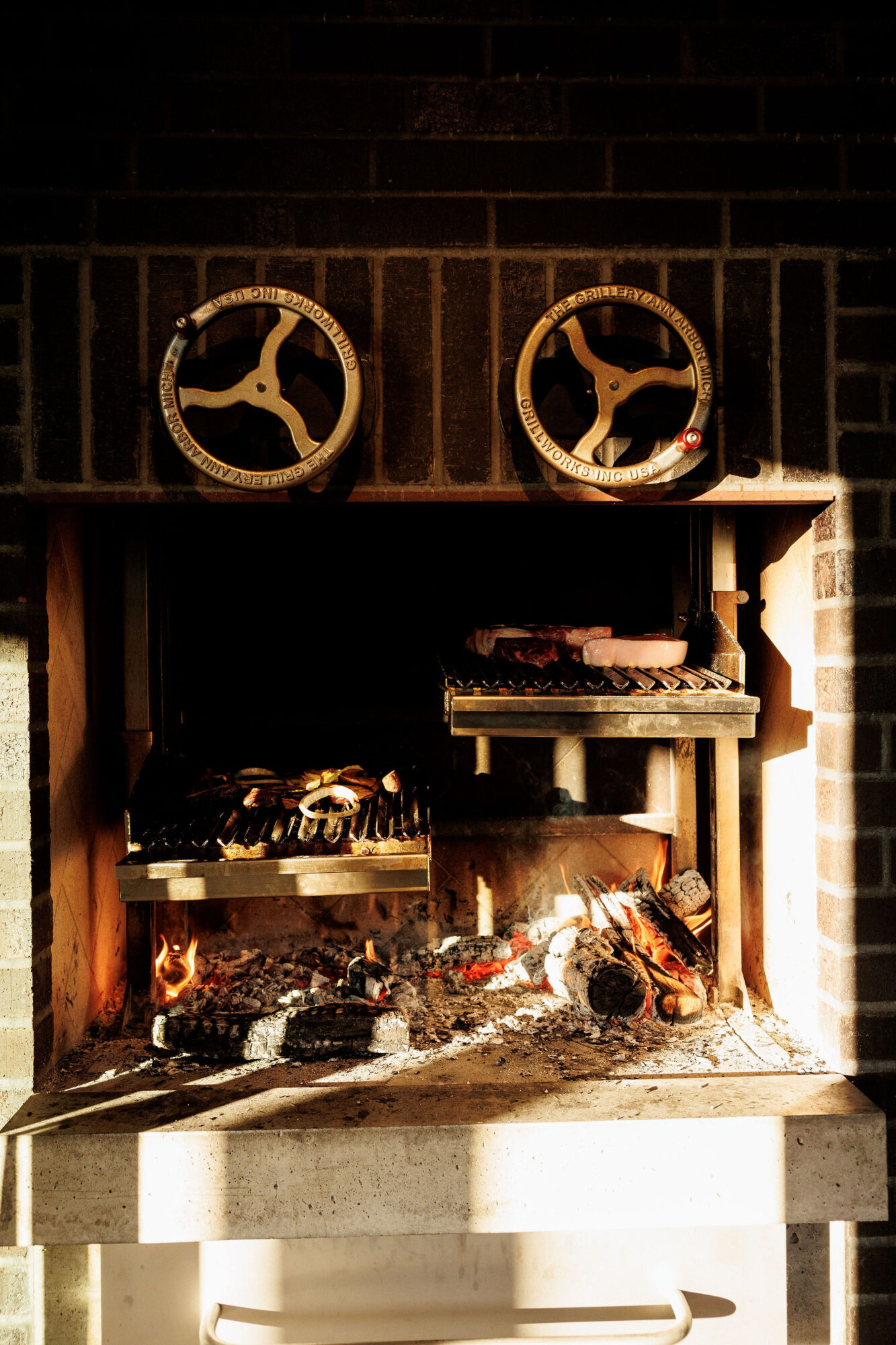 Seattle's best commercial food and hospitality photographer A wood-fired grill set into a brick wall, with burning logs and food cooking on metal grates. Captured by Seattles best commercial food lifestyle photographer, sunlight casts dramatic shadows across two large metal dials above the grill. captured by seattle's best food and lifestyle photographer Brooke Fitts