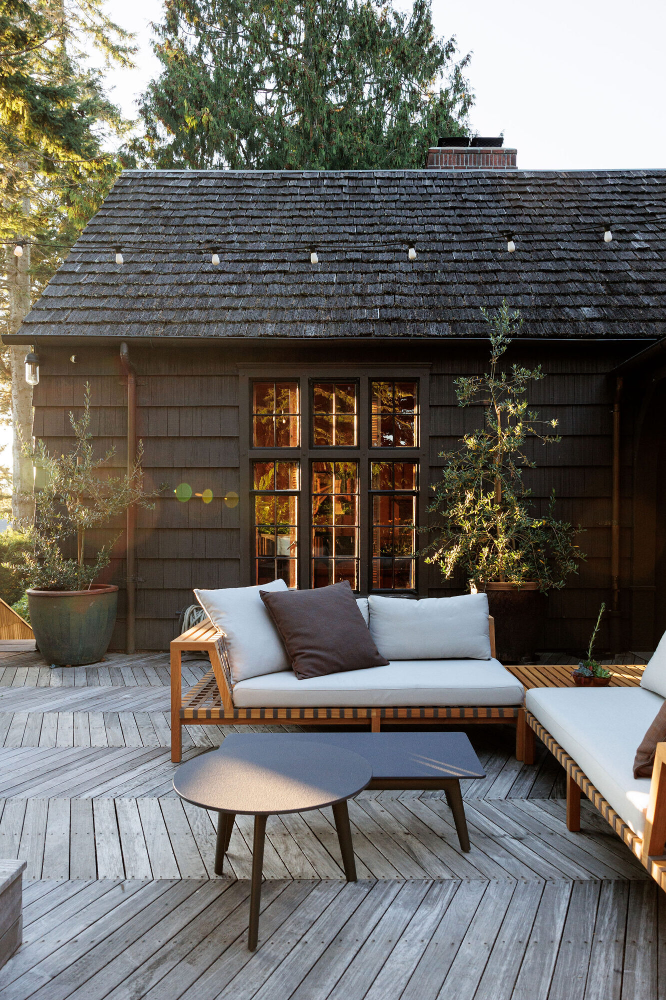 Seattle's best commercial food and hospitality photographer A cozy outdoor patio, styled by Seattles best commercial food lifestyle photographer, features a modern wooden sofa and coffee table on a deck. The rustic house backdrop boasts dark shingles, large windows, potted plants, and is surrounded by tall trees. captured by seattle's best food and lifestyle photographer Brooke Fitts