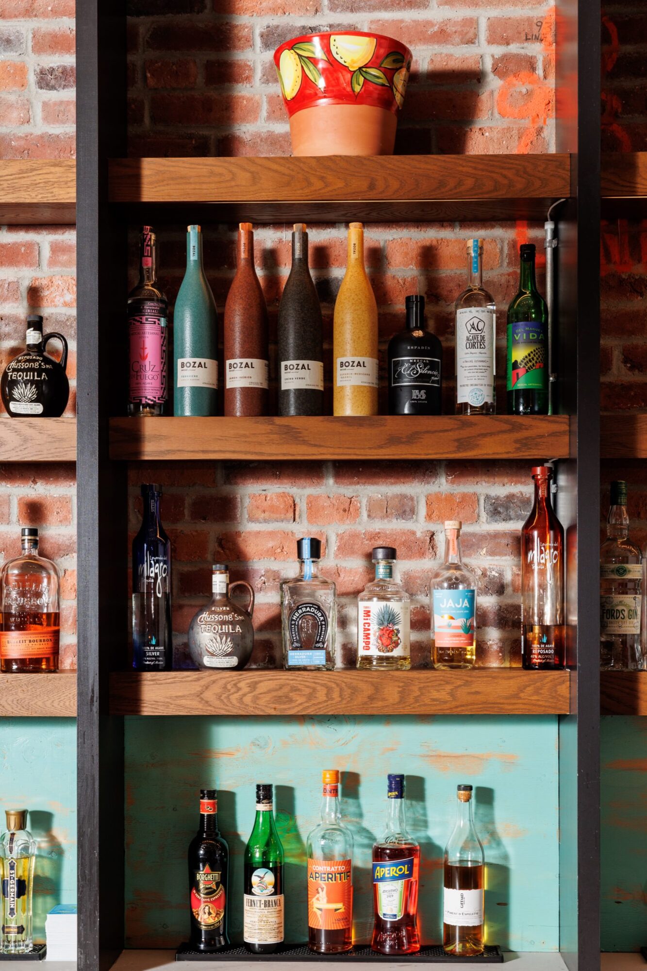 Seattle's best commercial food and hospitality photographer Shelves against a brick wall display various colorful liquor bottles, including mezcal, tequila, and other spirits. Captured by Seattles best commercial food lifestyle photographer, the setup has a rustic, vibrant bar atmosphere with a painted pot of lemons above. captured by seattle's best food and lifestyle photographer Brooke Fitts