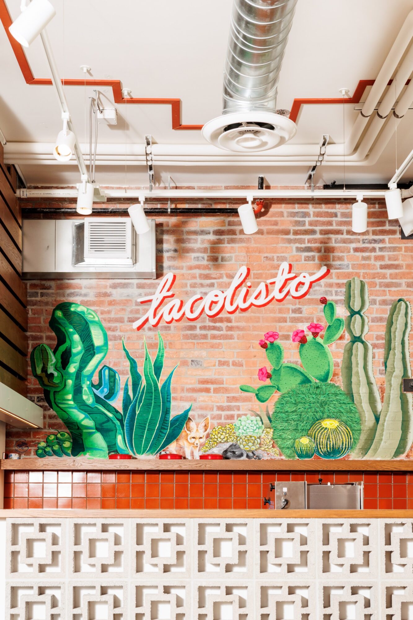 Seattle's best commercial food and hospitality photographer A brightly lit restaurant wall features a neon “tacolisto” sign above colorful painted cacti and desert plants against exposed brick, captured by Seattles best commercial food lifestyle photographer, with red tiles and decorative white blocks in the foreground. captured by seattle's best food and lifestyle photographer Brooke Fitts