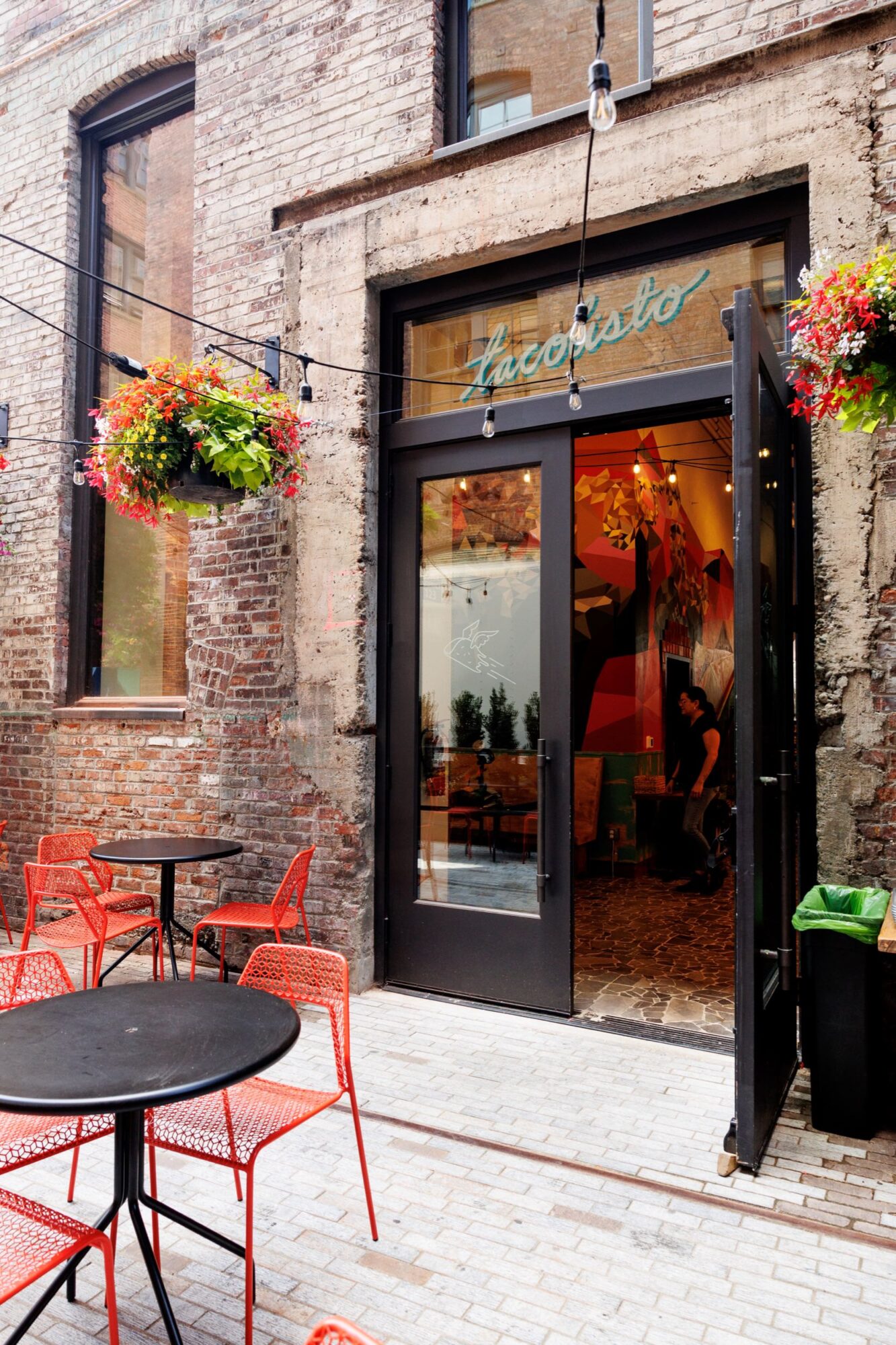 Seattle's best commercial food and hospitality photographer A brick-walled patio with red chairs and black tables sits outside a restaurant called Tacopisto. Captured by Seattles best commercial food lifestyle photographer, the inviting outdoor space features hanging flower baskets and string lights. captured by seattle's best food and lifestyle photographer Brooke Fitts