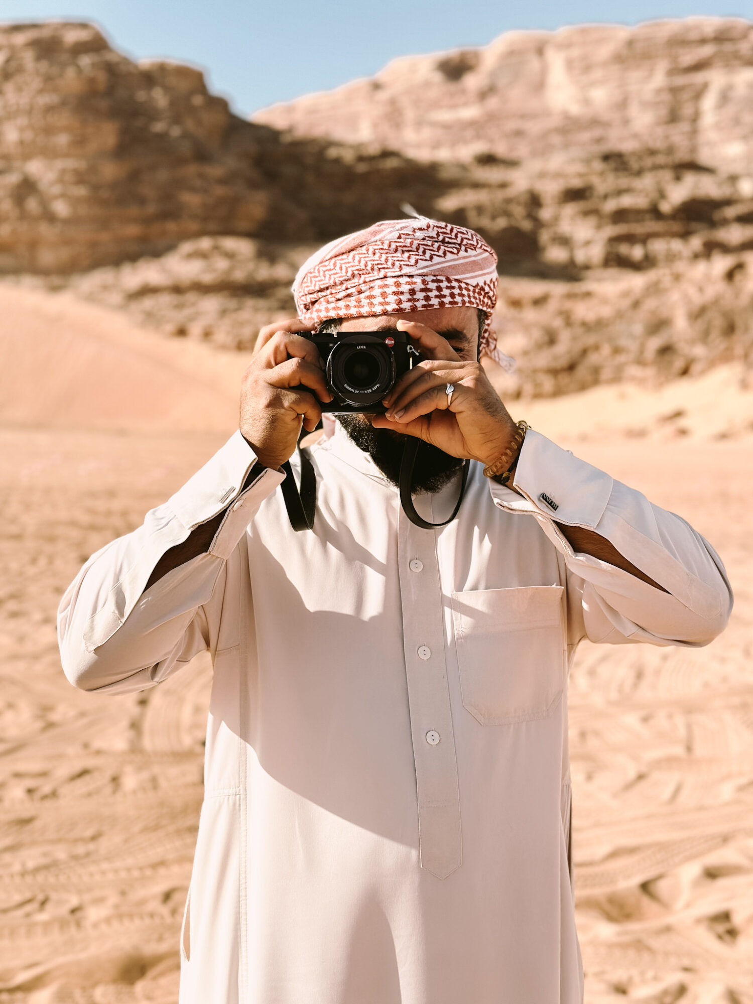 Seattle's best commercial food and hospitality photographer A person wearing a white thobe and patterned headscarf stands in a sandy desert landscape, holding a camera up to their face and taking a photo, capturing the scene like Seattles best commercial food lifestyle photographer, with rocky hills in the background. captured by seattle's best food and lifestyle photographer Brooke Fitts