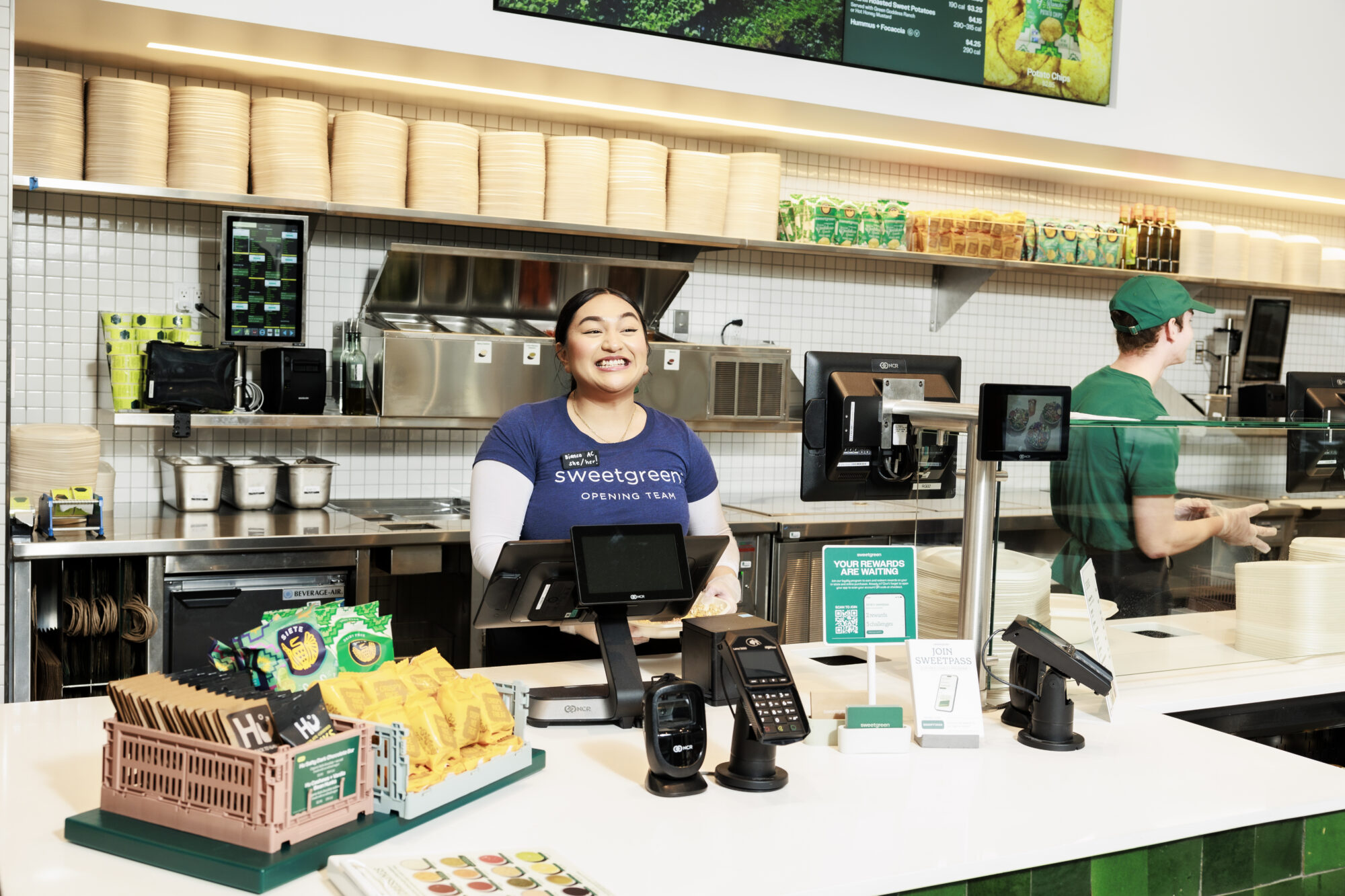 Seattle's best commercial food and hospitality photographer A smiling employee stands behind the counter at a Sweetgreen restaurant, with salads, menu displays, and fresh ingredients visible in the bright, modern interior—captured by Seattles best commercial food lifestyle photographer. Another worker prepares food in the background. captured by seattle's best food and lifestyle photographer Brooke Fitts