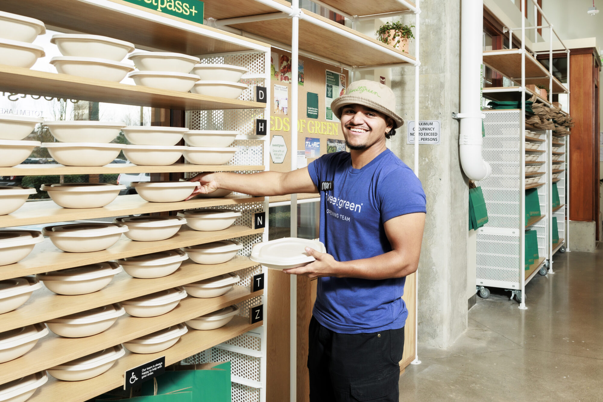 Seattle's best commercial food and hospitality photographer A smiling person in a blue shirt and bucket hat holds a bowl while placing or retrieving it from a shelf lined with stacked bowls in a bright, modern eatery—captured by Seattles best commercial food lifestyle photographer. captured by seattle's best food and lifestyle photographer Brooke Fitts