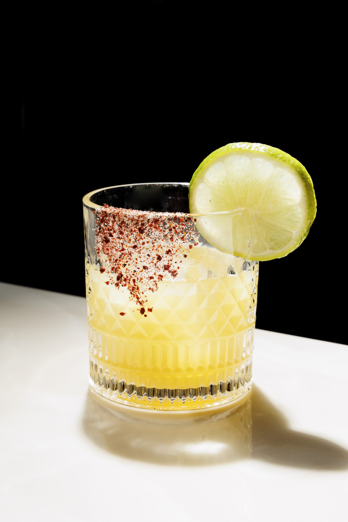 Seattle's best commercial food and hospitality photographer A short glass filled with a yellow cocktail, rimmed with chili powder and garnished with a fresh lime wheel, is artfully captured by Seattles best commercial food lifestyle photographer against a black background on a white surface. captured by seattle's best food and lifestyle photographer Brooke Fitts