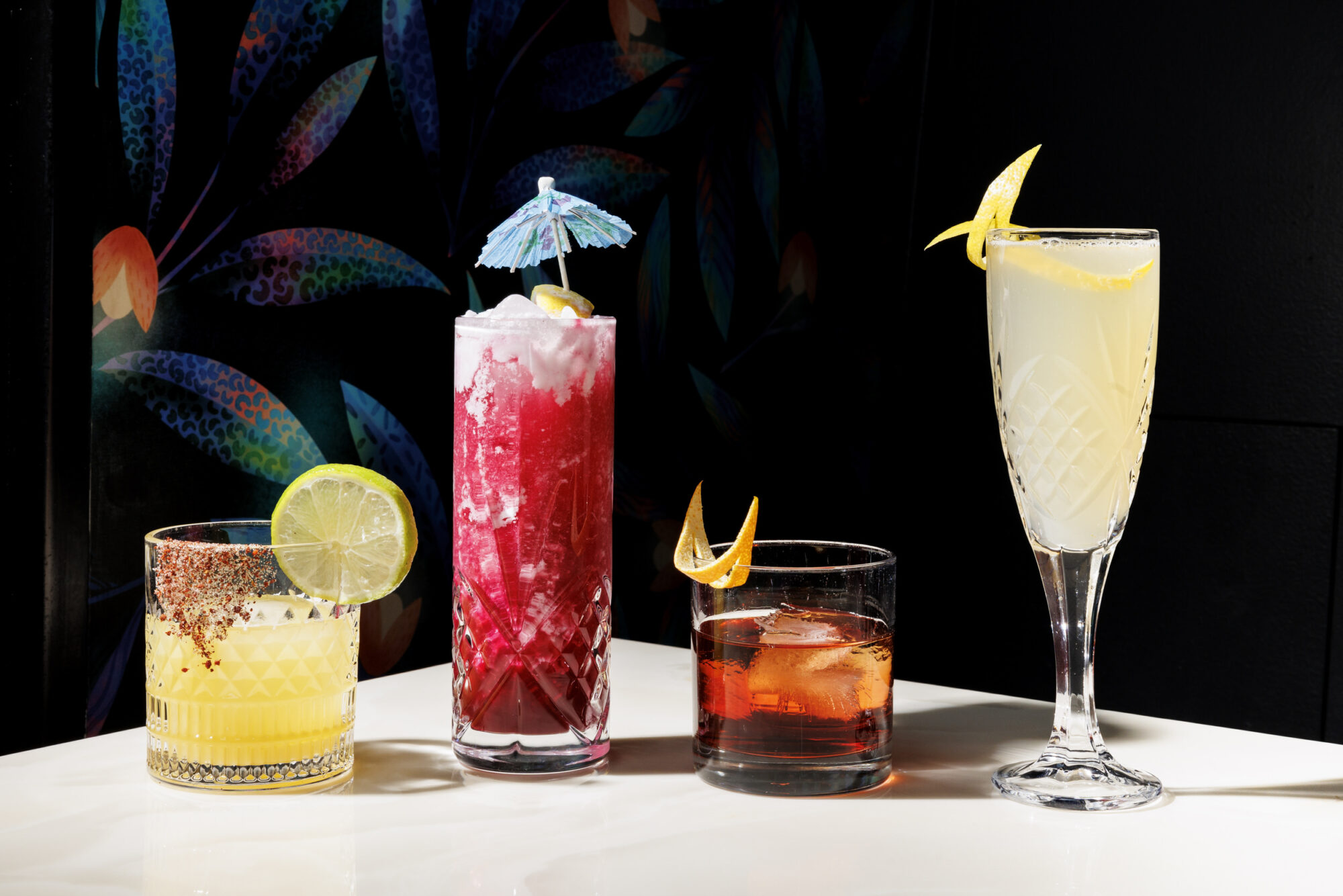 Seattle's best commercial food and hospitality photographer Four colorful cocktails on a white surface, artfully captured by Seattles best commercial food lifestyle photographer: a yellow drink with lime, a tall red drink with crushed ice and blue umbrella, an amber drink with orange twist, and a pale flute with lemon garnish. captured by seattle's best food and lifestyle photographer Brooke Fitts