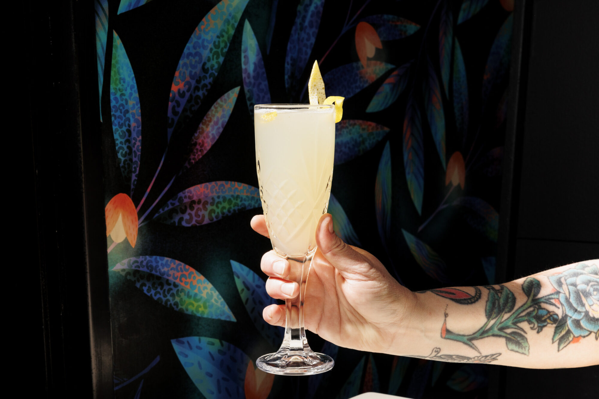 Seattle's best commercial food and hospitality photographer A tattooed hand holds a champagne flute filled with a pale yellow drink garnished with a lemon twist, against a colorful, leafy patterned background—captured by Seattles best commercial food lifestyle photographer. captured by seattle's best food and lifestyle photographer Brooke Fitts