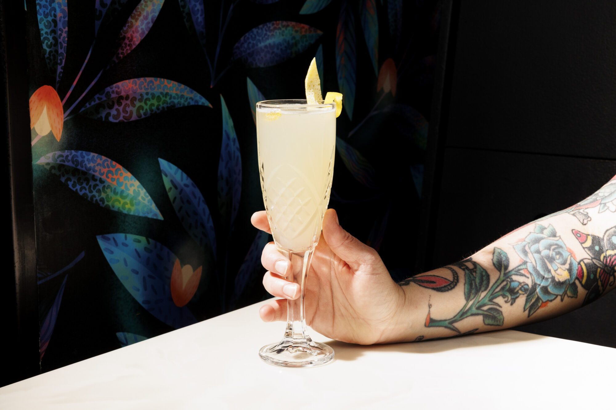 Seattle's best commercial food and hospitality photographer A tattooed arm holds a tall, elegant glass of pale yellow cocktail garnished with a lemon twist, captured by Seattles best commercial food lifestyle photographer against a dark, floral-patterned background. captured by seattle's best food and lifestyle photographer Brooke Fitts
