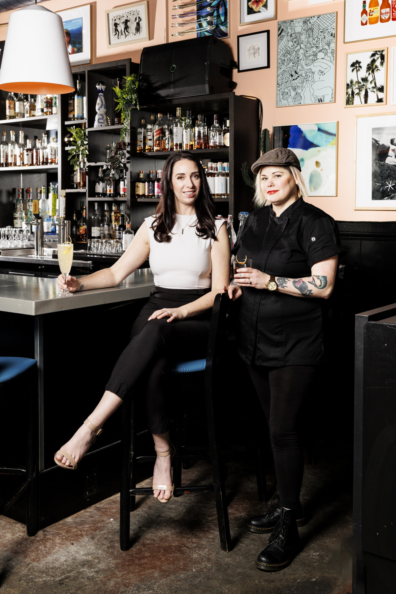 Seattle's best commercial food and hospitality photographer Two women pose in a stylish bar—one seated on a stool with a drink, the other standing in black attire and hat. Captured by Seattles best commercial food lifestyle photographer, bar shelves and framed art decorate the vibrant background. captured by seattle's best food and lifestyle photographer Brooke Fitts
