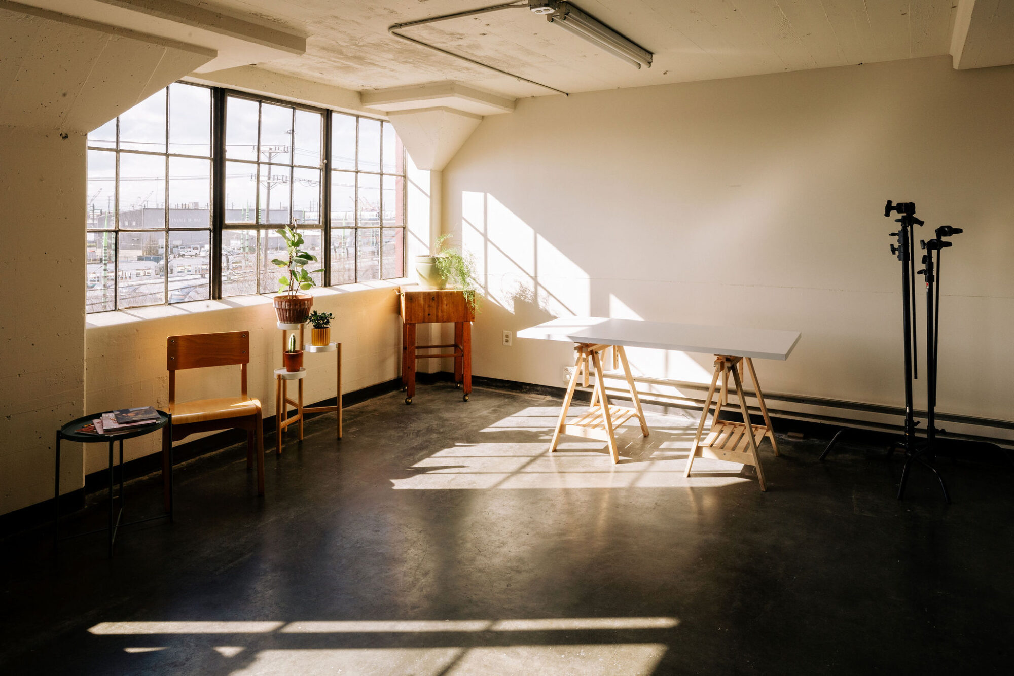 Seattle's best commercial food and hospitality photographer A bright, minimalist room with large windows, a white desk on wooden sawhorses, a few wooden chairs, two potted plants, and two camera tripods—perfect for Seattles best commercial food lifestyle photographer. Sunlight streams in, casting shadows on the floor. captured by seattle's best food and lifestyle photographer Brooke Fitts