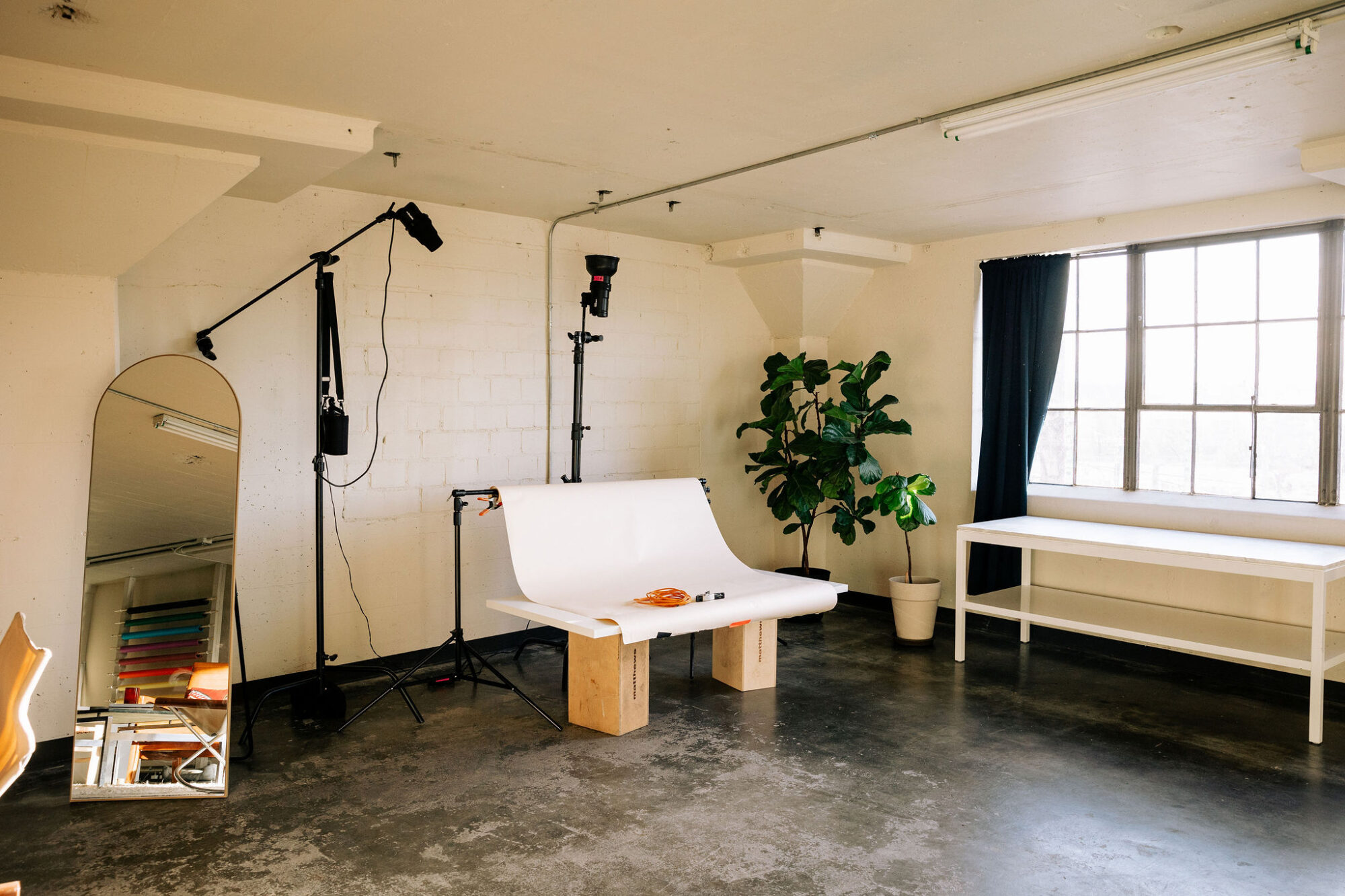 Seattle's best commercial food and hospitality photographer A photo studio set up by Seattles best commercial food lifestyle photographer, featuring a white backdrop on wooden blocks, professional lighting, a large potted plant, a white table, mirror, and daylight streaming through dark-curtained windows. captured by seattle's best food and lifestyle photographer Brooke Fitts