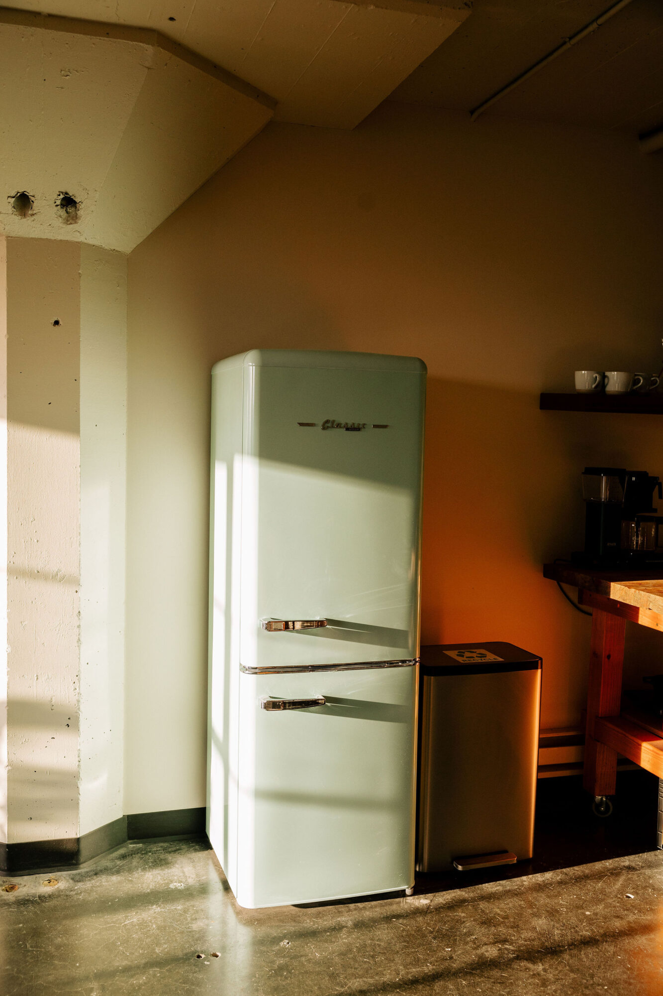 Seattle's best commercial food and hospitality photographer A vintage-style light green refrigerator stands next to a stainless steel trash bin in a sunlit kitchen, beautifully captured by Seattles best commercial food lifestyle photographer. Beige walls and a wooden shelf with mugs and coffee supplies complete the scene. captured by seattle's best food and lifestyle photographer Brooke Fitts