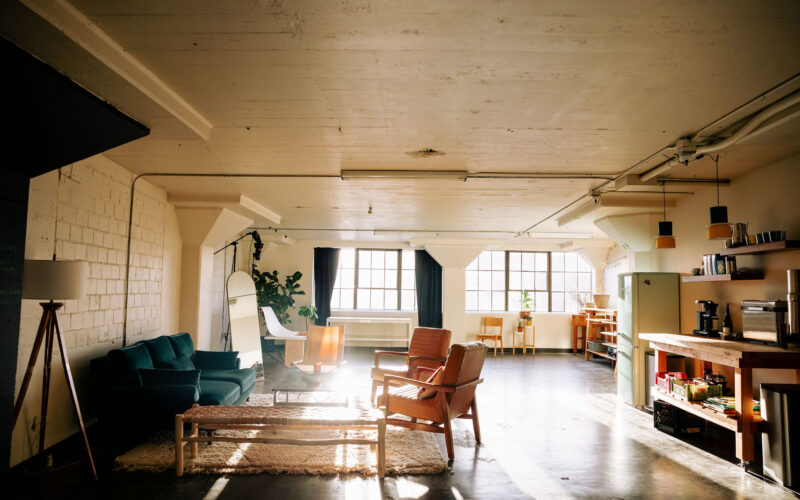 Seattle's best commercial food and hospitality photographer Sunlit loft apartment with a green sofa, armchairs, a coffee table, and a cozy rug. Captured by Seattles best commercial food lifestyle photographer, the space features large windows, kitchen shelves, and vibrant plants and decor throughout. captured by seattle's best food and lifestyle photographer Brooke Fitts