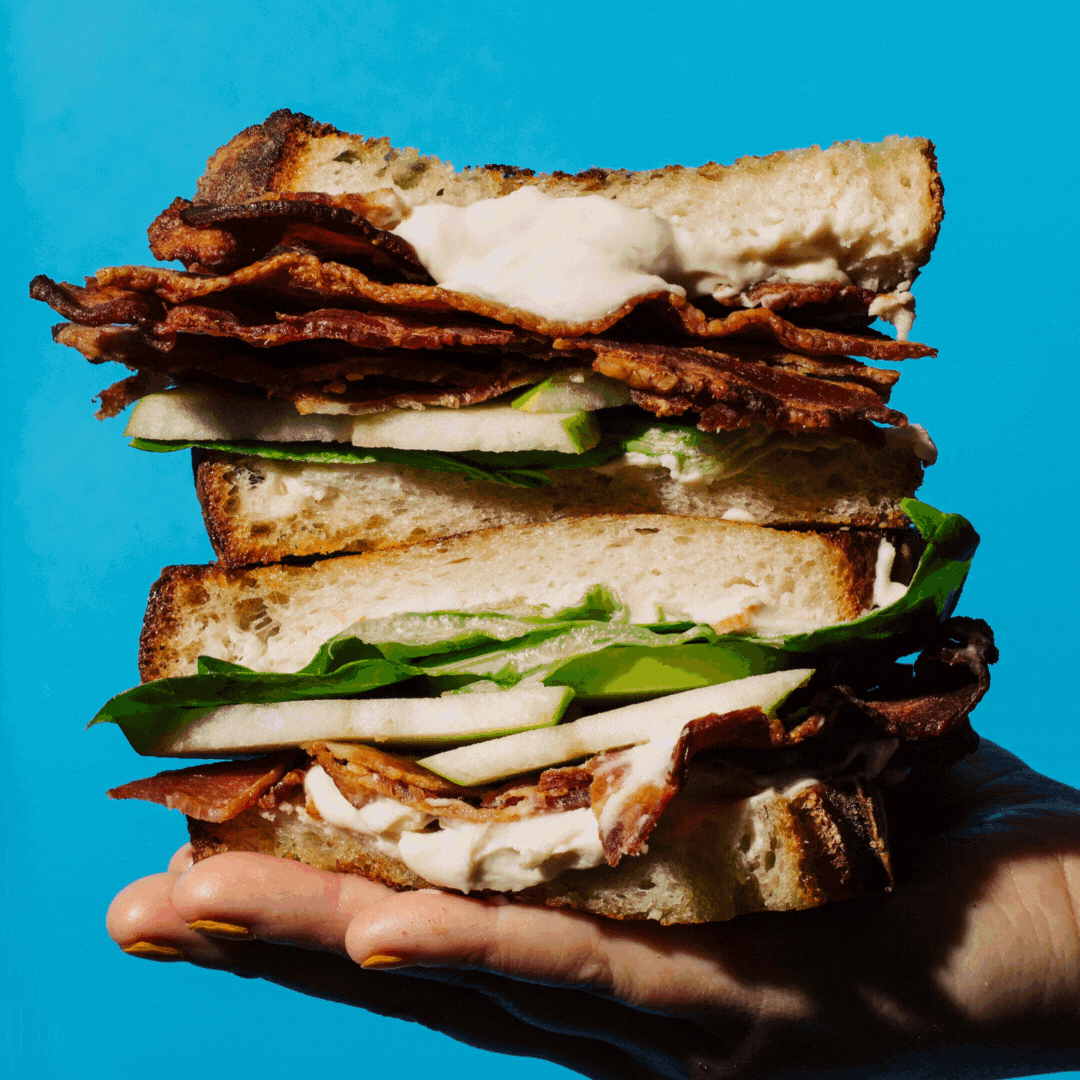 Seattle's best commercial food and hospitality photographer A hand holds a thick sandwich packed with crispy bacon, lettuce, apple slices, and creamy sauce between rustic bread, captured against a bright blue background by Seattles best commercial food lifestyle photographer. captured by seattle's best food and lifestyle photographer Brooke Fitts