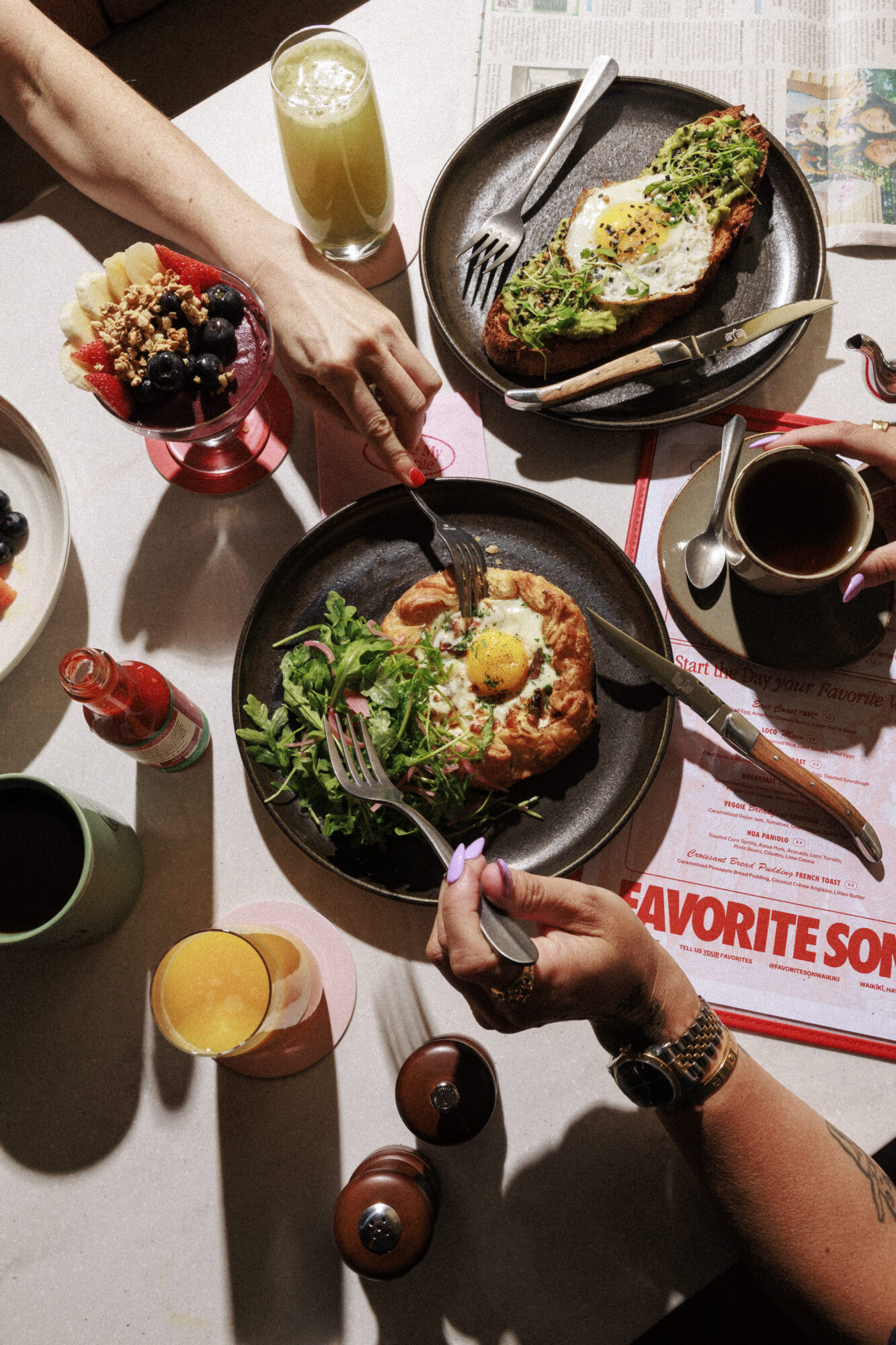 Seattle's best commercial food and hospitality photographer A breakfast spread captured by Seattles best commercial food lifestyle photographer features avocado toast topped with egg, a pastry with greens and egg, fruit parfait, coffee, orange juice, and hands reaching for food on a sunlit table. captured by seattle's best food and lifestyle photographer Brooke Fitts