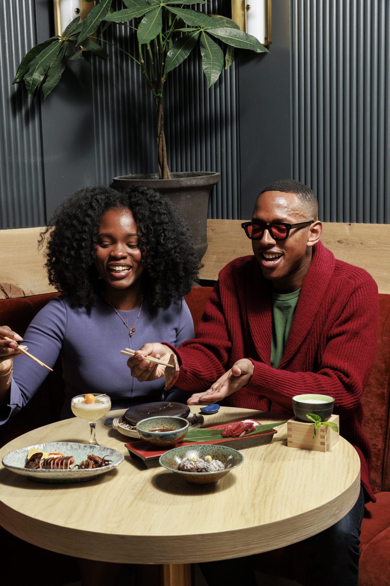 Seattle's best commercial food and hospitality photographer Two people sit at a round table enjoying a meal together, smiling and using chopsticks. Captured by Seattles best commercial food lifestyle photographer, various dishes and drinks are arranged before them, with a potted plant in the background. captured by seattle's best food and lifestyle photographer Brooke Fitts