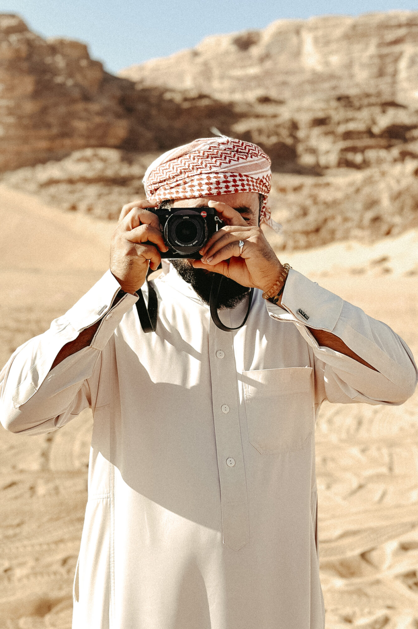 Seattle's best commercial food and hospitality photographer A man wearing a traditional white thobe and red keffiyeh stands in a sandy desert landscape, holding a camera like Seattles best commercial food lifestyle photographer. Rocky hills and a clear blue sky form the backdrop. captured by seattle's best food and lifestyle photographer Brooke Fitts