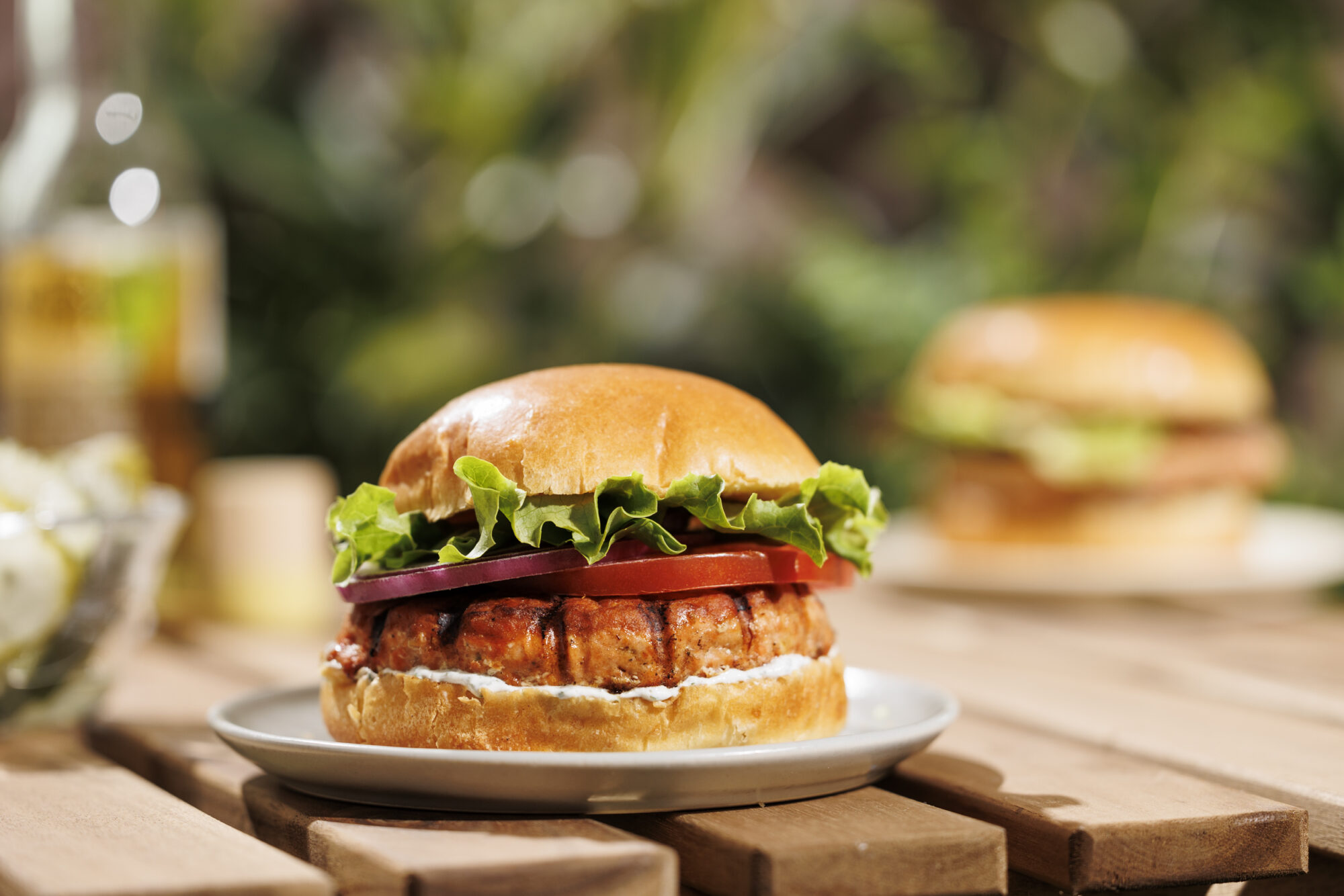 Seattle's best commercial food and hospitality photographer A close-up of a burger with lettuce, tomato, and a patty in a shiny bun, styled by Seattles best commercial food lifestyle photographer, served on a white plate on a wooden table with another burger blurred in the background. captured by seattle's best food and lifestyle photographer Brooke Fitts