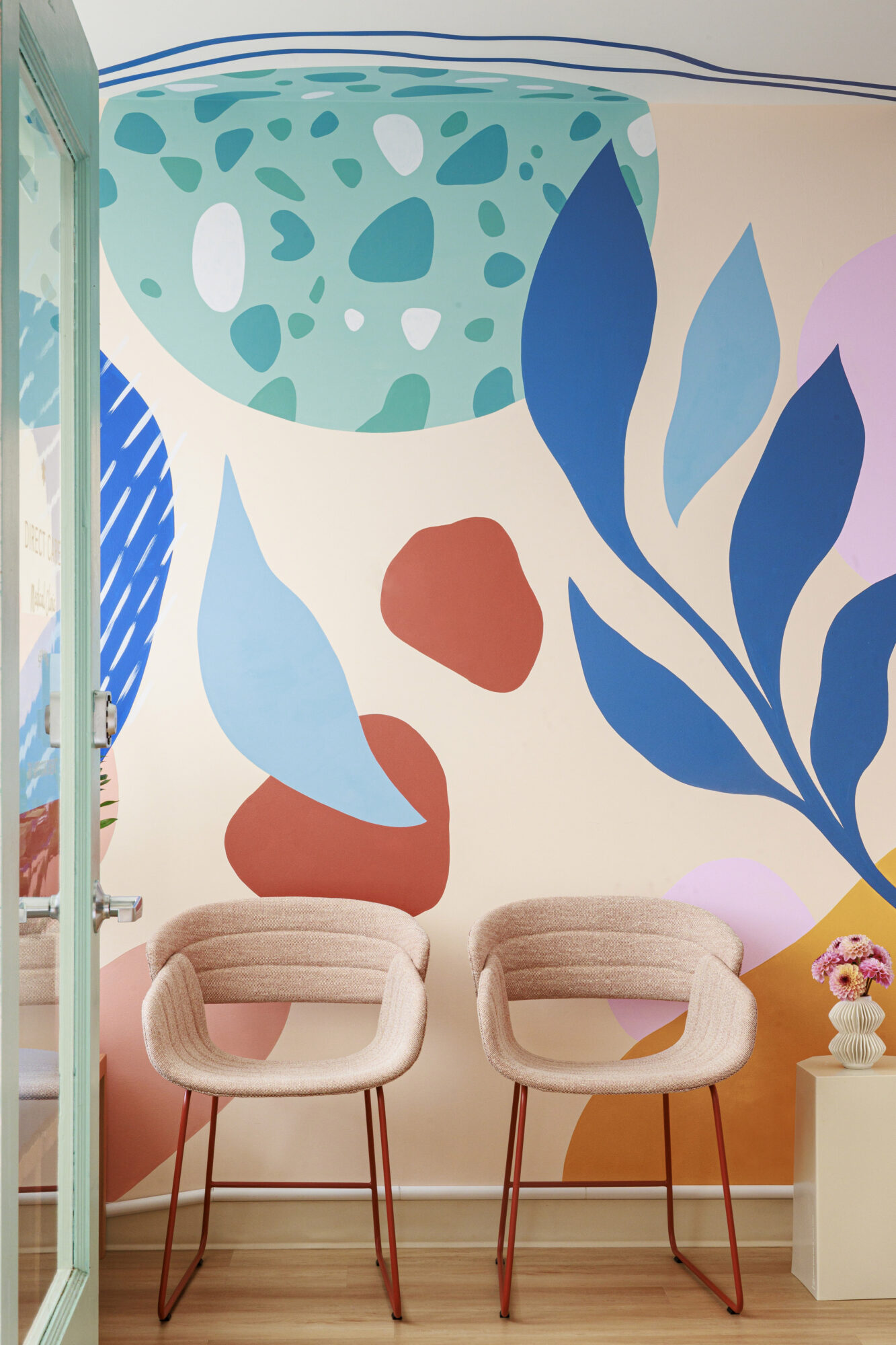 Seattle's best commercial food and hospitality photographer Two pink chairs with red legs sit against a vibrant wall mural of abstract shapes and bold leaves. Styled by Seattle’s best commercial food lifestyle photographer, a small table with a vase of flowers completes the inviting scene. captured by seattle's best food and lifestyle photographer Brooke Fitts