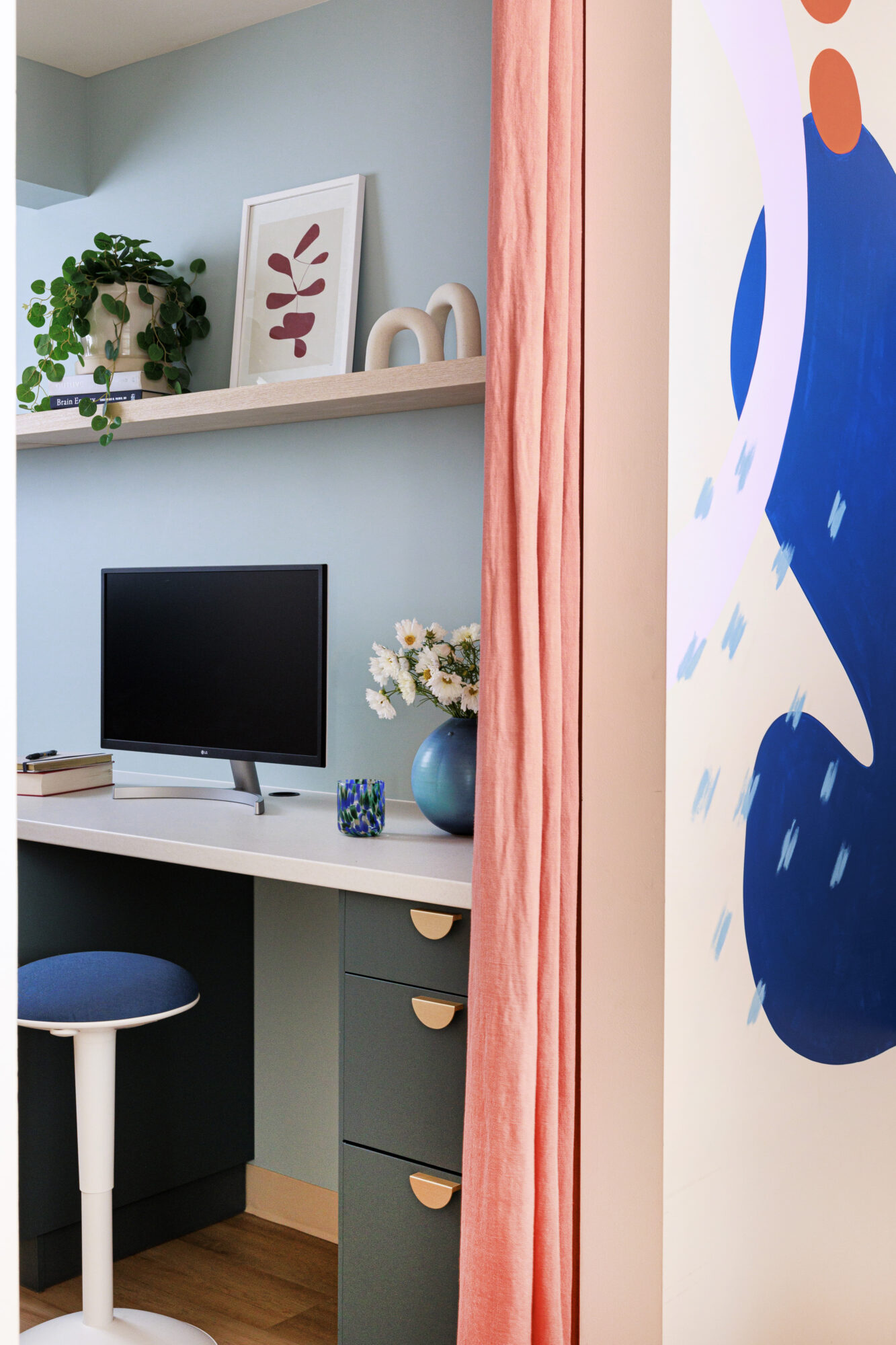 Seattle's best commercial food and hospitality photographer A modern home office nook with a computer, chair, books, potted plant, art, and a vase of flowers on a desk—partially enclosed by a pink curtain and a colorful abstract wall mural—captured by Seattles best commercial food lifestyle photographer. captured by seattle's best food and lifestyle photographer Brooke Fitts
