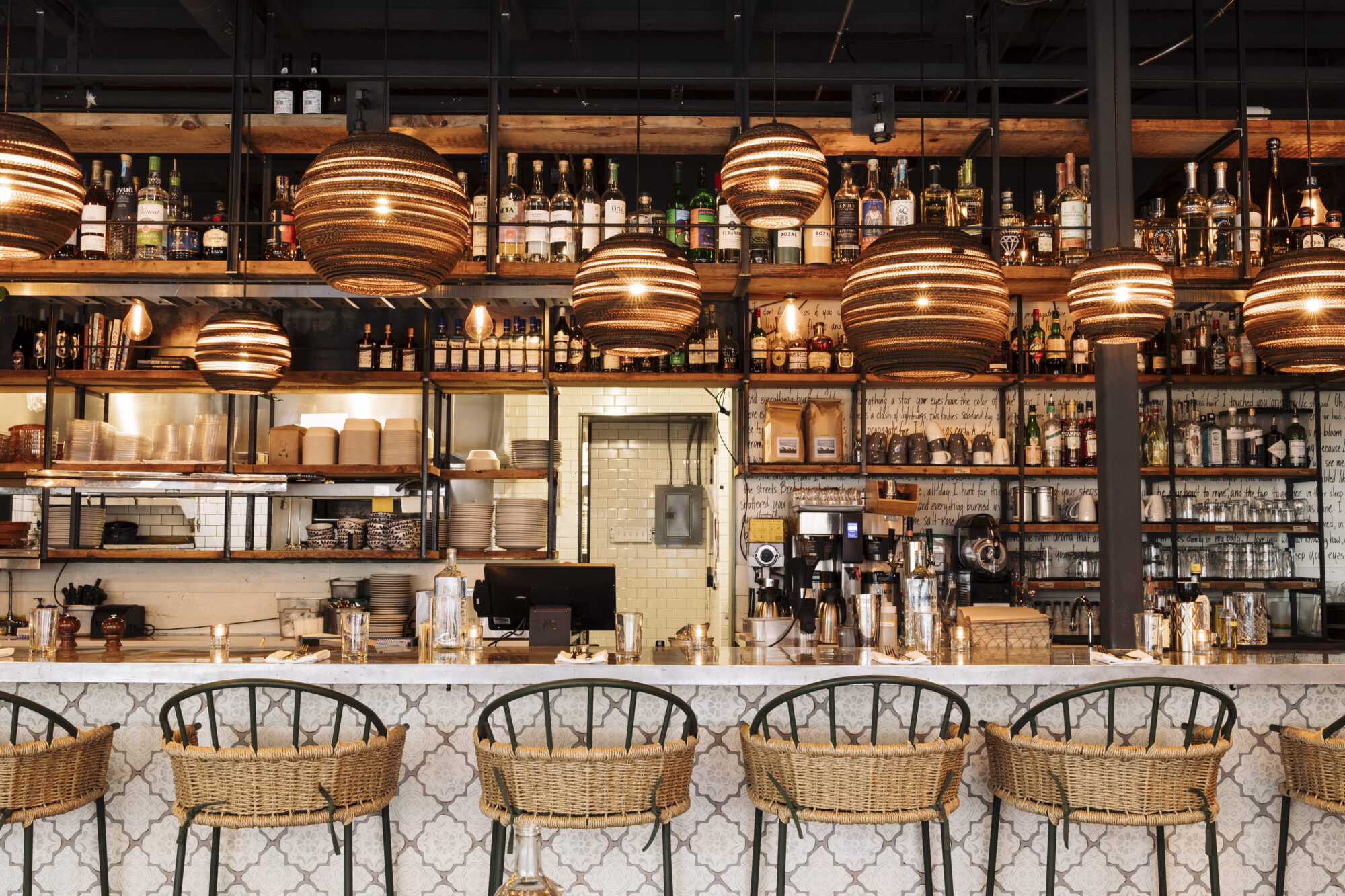 Seattle's best commercial food and hospitality photographer A stylish bar with woven pendant lights, wicker bar stools, and shelves filled with bottles and glassware behind a white marble counter in a modern, cozy setting—captured by a Seattle commercial interior design photographer for restaurants. captured by seattle's best food and lifestyle photographer Brooke Fitts