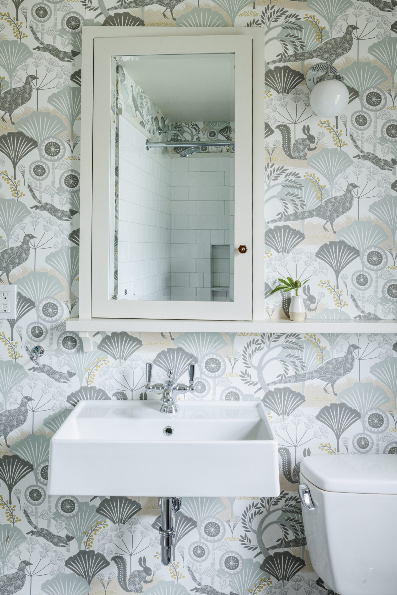 Seattle's best commercial interiors and hospitality photographer A bathroom with patterned wallpaper, a rectangular mirror cabinet above a white sink, a small plant on a shelf, and a white wall light—captured by a Seattle commercial interior design photographer for restaurants and hotels. captured by seattle's best food and lifestyle photographer Brooke Fitts
