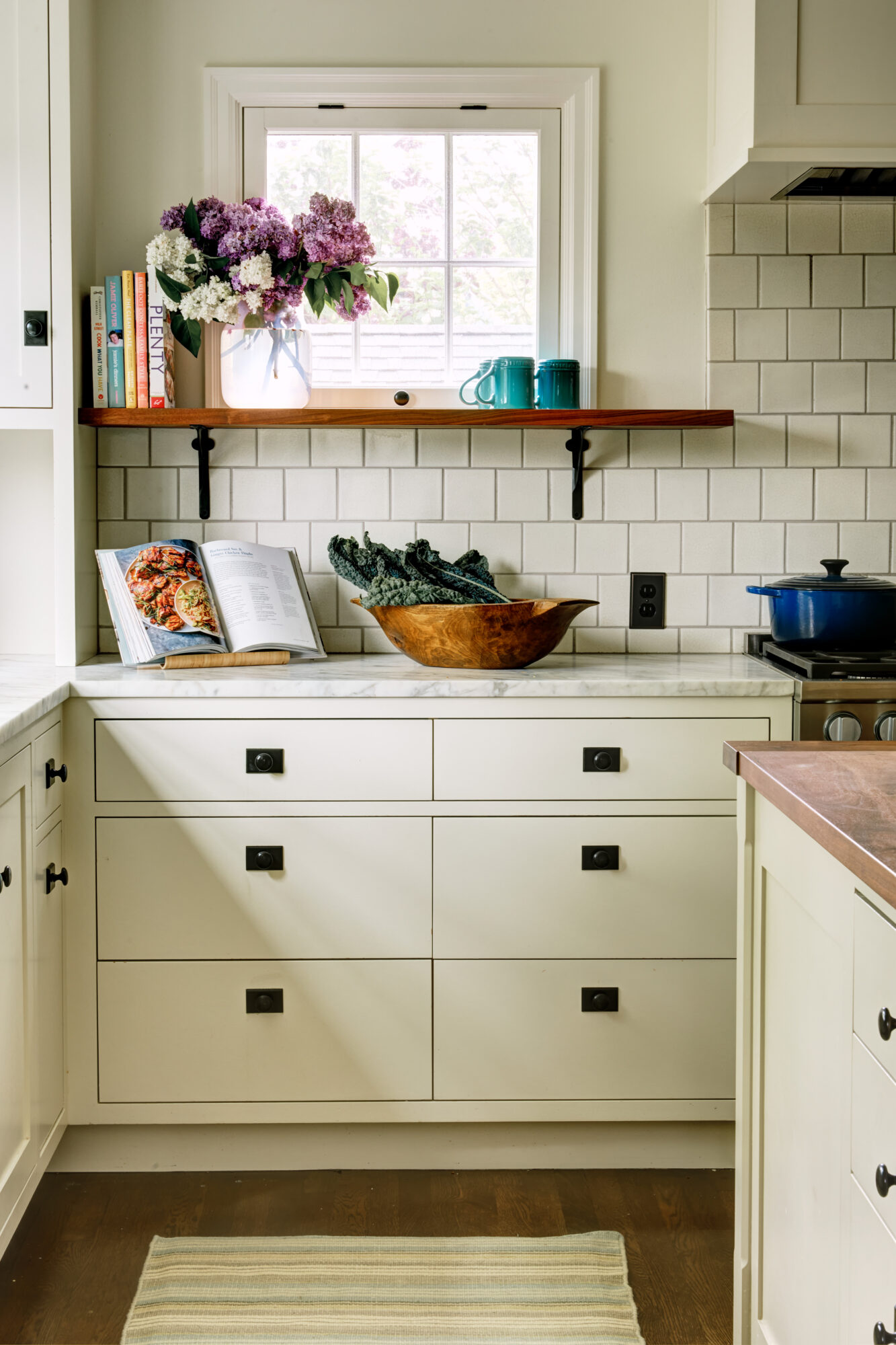 Seattle's best commercial interiors and hospitality photographer A bright kitchen with white cabinets, a tile backsplash, and a striped rug. Captured by a Seattle commercial interior design photographer for restaurants and hotels, the space features a wooden bowl, open cookbook, and a shelf with flowers beneath a small window. captured by seattle's best food and lifestyle photographer Brooke Fitts