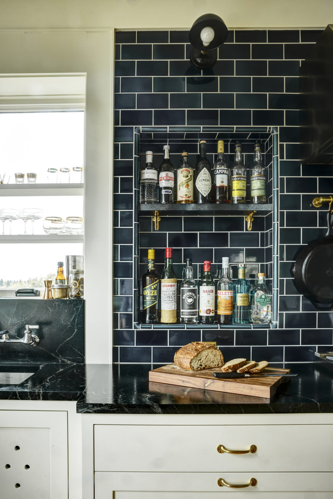 Seattle's best commercial interiors and hospitality photographer A kitchen with a dark subway tile backsplash, a built-in shelf holding various bottles, a black countertop with a cutting board displaying sliced bread, and a window with glassware—captured by a Seattle commercial interior design photographer. captured by seattle's best food and lifestyle photographer Brooke Fitts
