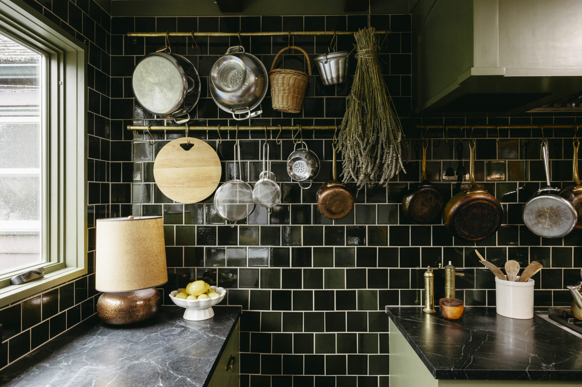Seattle's best commercial interiors and hospitality photographer A cozy kitchen with glossy dark green tiled walls, hanging pots, pans, and baskets is beautifully captured by a Seattle commercial interior design photographer for restaurants and hotels; the space features dried herbs, a round cutting board, and lemons on black marble. captured by seattle's best food and lifestyle photographer Brooke Fitts