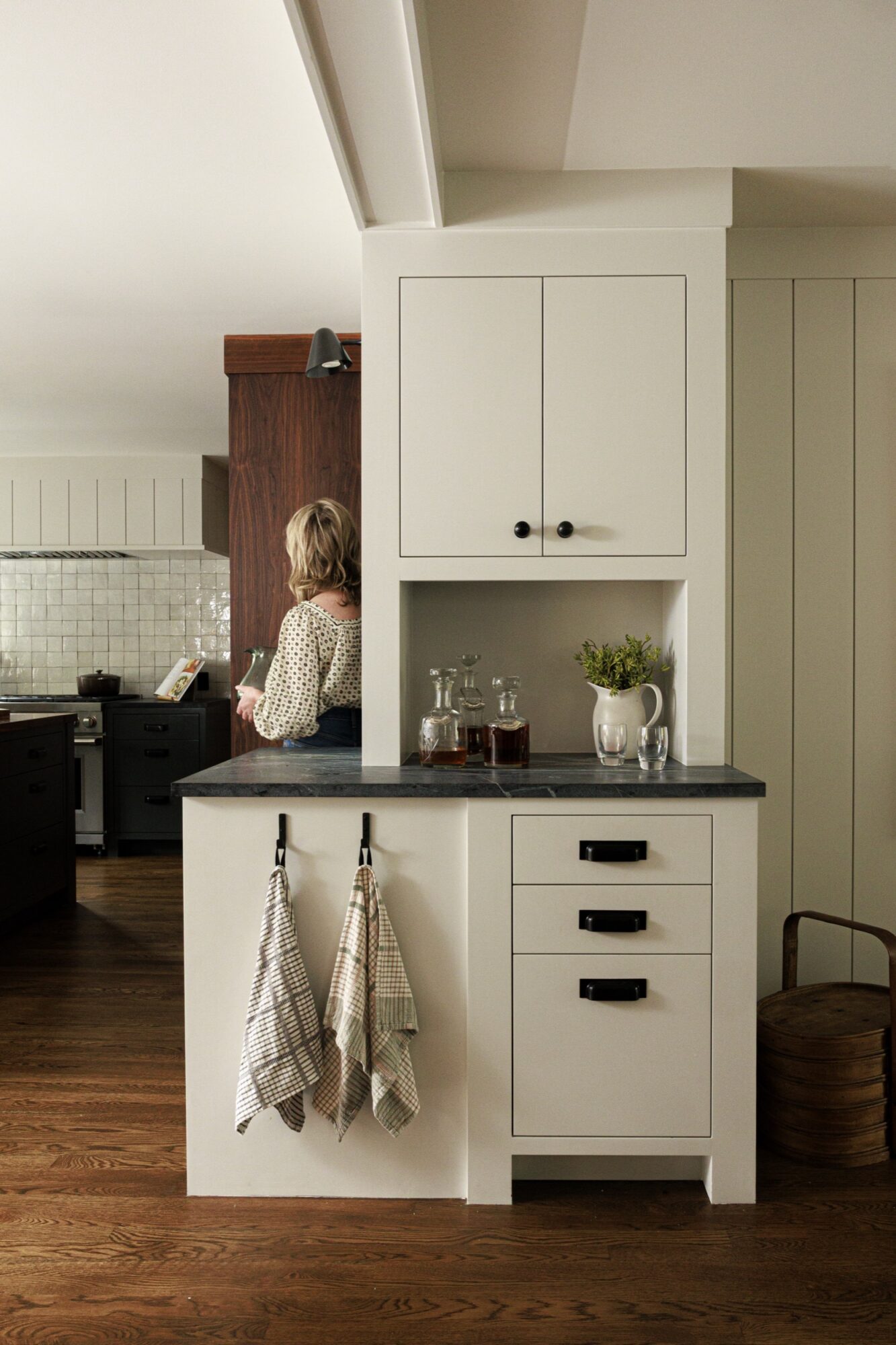 Seattle's best commercial food and hospitality photographer A woman stands in a modern kitchen with white cabinets, dark countertops, and wooden floors. Captured by a Seattle commercial interior design photographer for restaurants and hotels, the space features towels on hooks and glass bottles with greenery on the counter. captured by seattle's best food and lifestyle photographer Brooke Fitts