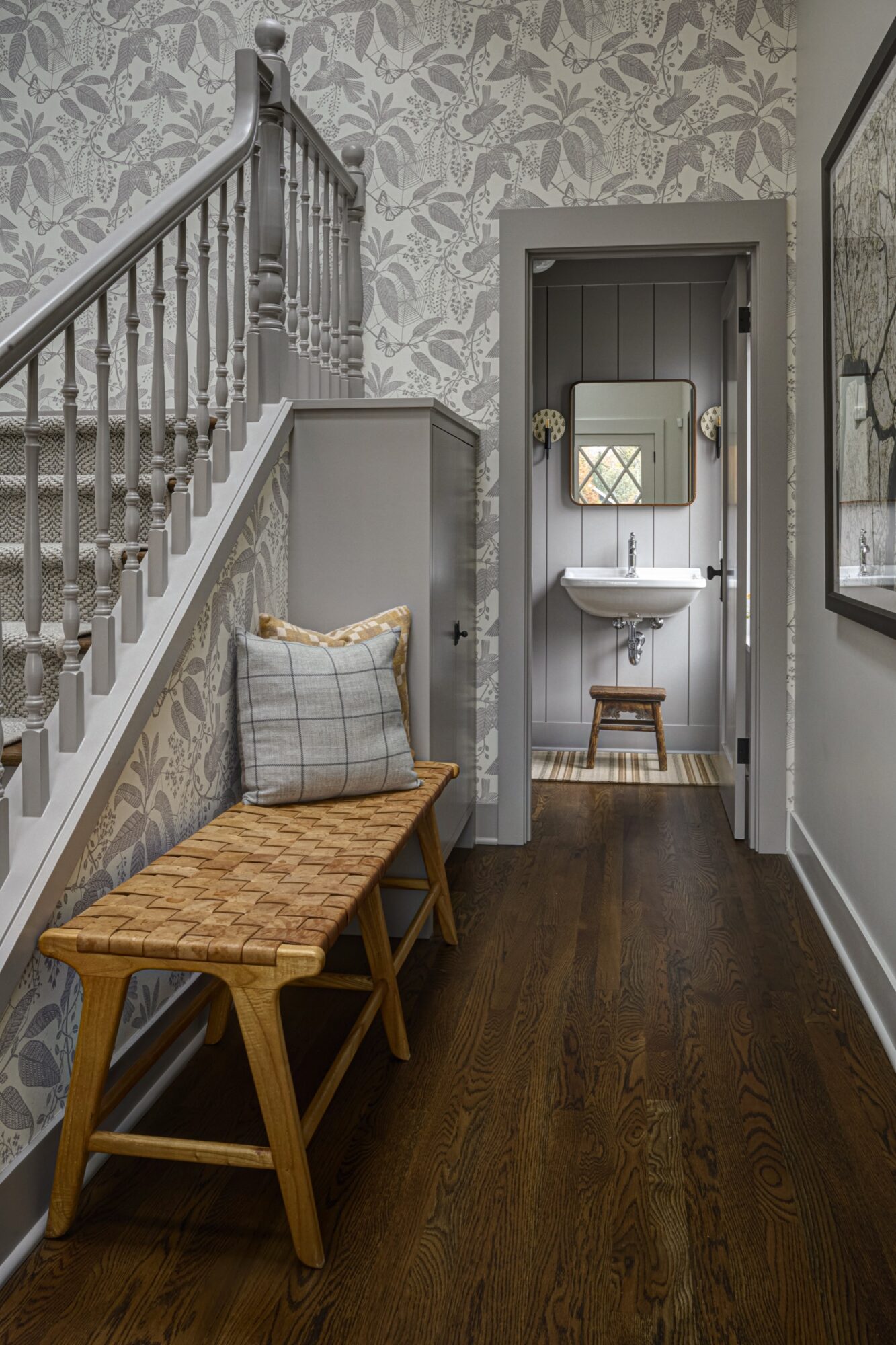 Seattle's best commercial food and hospitality photographer A hallway with floral wallpaper, wooden floor, and a woven bench with plaid and tan pillows under a staircase is beautifully captured by a Seattle commercial interior design photographer for restaurants and hotels, offering a glimpse into a gray bathroom. captured by seattle's best food and lifestyle photographer Brooke Fitts
