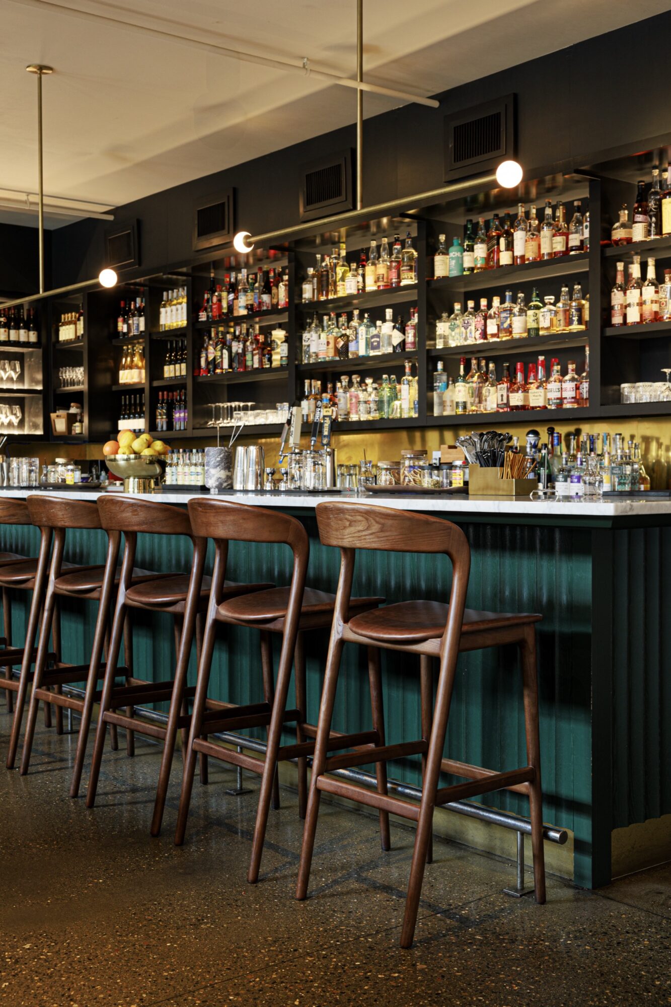 Seattle's best commercial food and hospitality photographer A modern bar with a long counter, four wooden bar stools, and shelves stocked with bottles of liquor and glasses. Warm lighting and an inviting atmosphere are captured by a Seattle commercial interior design photographer for restaurants and hotels. captured by seattle's best food and lifestyle photographer Brooke Fitts