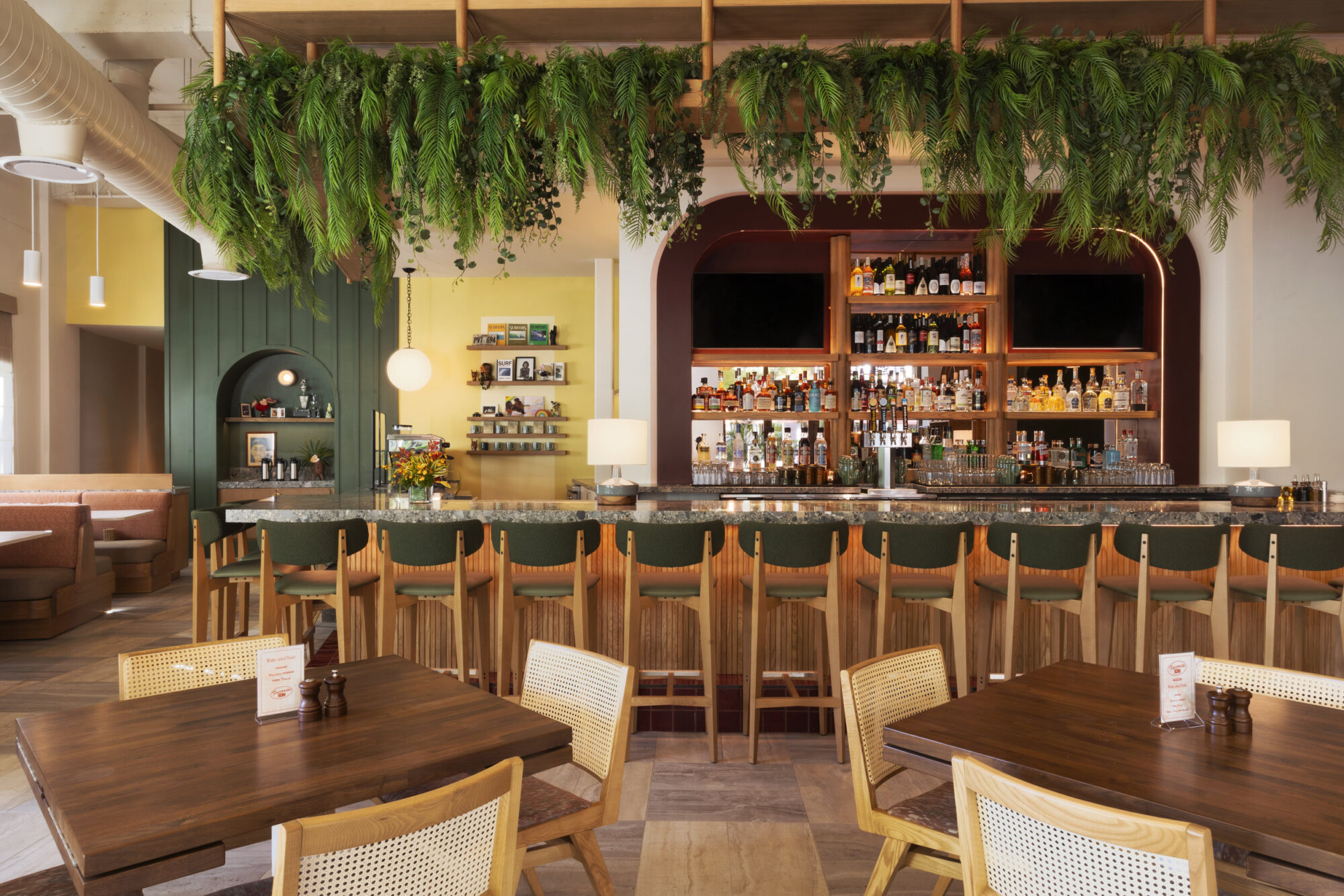 Seattle's best commercial food and hospitality photographer A modern bar with wooden chairs and tables, a green countertop, and shelves stocked with bottles behind the bar. Captured by a Seattle commercial interior design photographer for restaurants and hotels, green plants hang above, adding a fresh touch to the bright, stylish space. captured by seattle's best food and lifestyle photographer Brooke Fitts