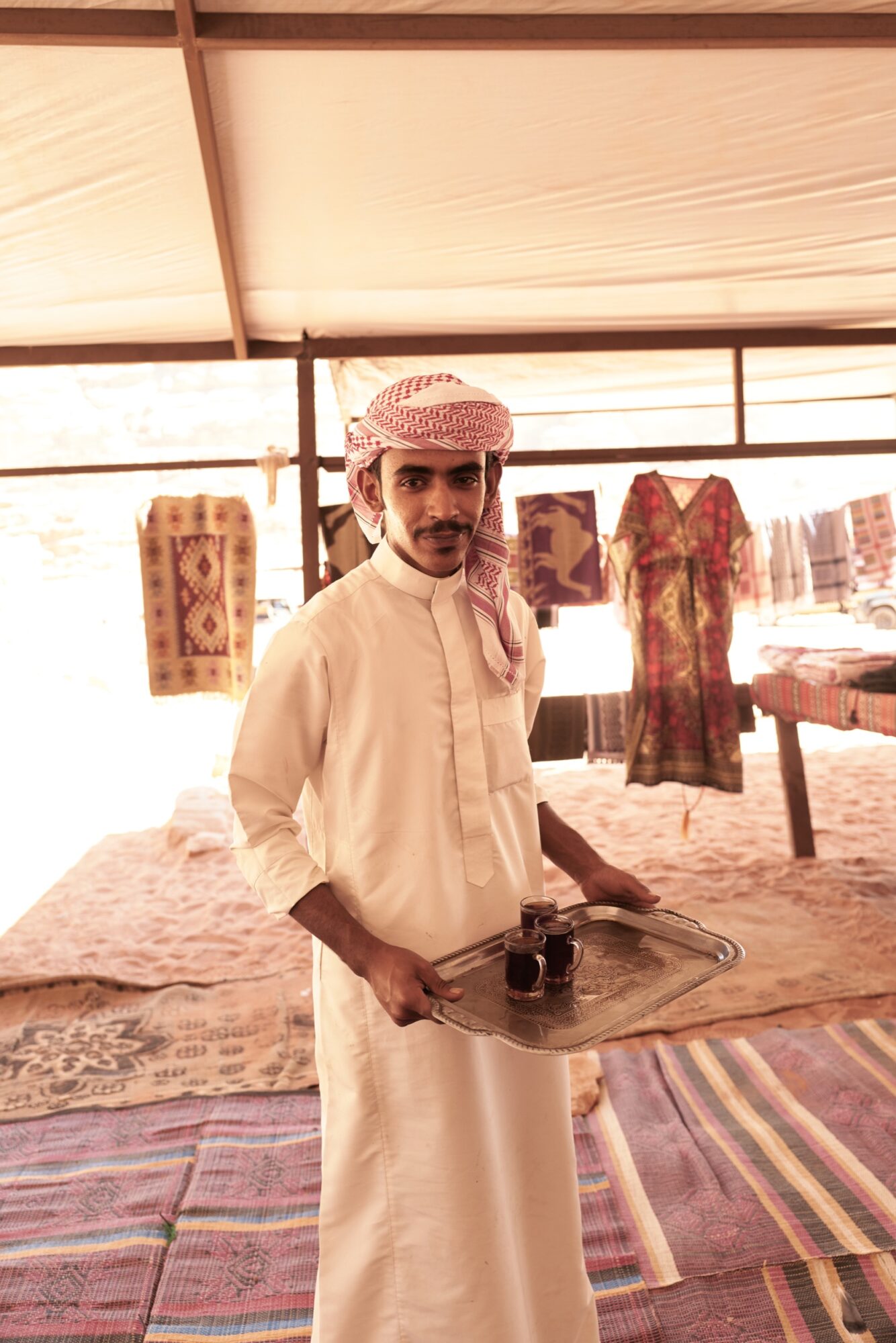 Seattle's best commercial food and hospitality photographer A man wearing traditional Middle Eastern attire and a red-and-white headscarf stands in a tent, holding a tray with two cups of coffee. Colorful rugs and textiles hang and lay around him. captured by seattle's best food and lifestyle photographer Brooke Fitts