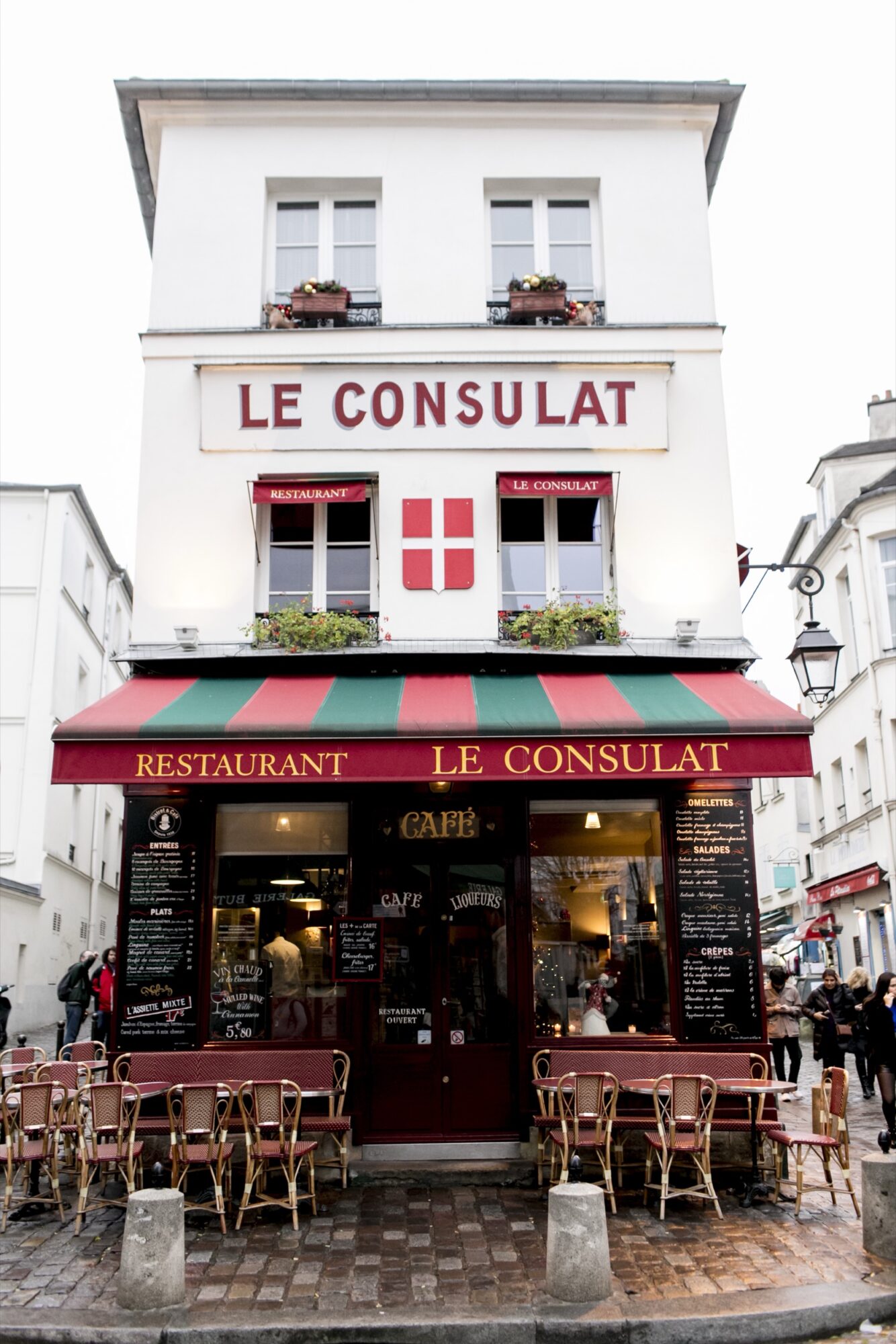 Seattle's best commercial food and hospitality photographer A charming Parisian café named Le Consulat with a red and green striped awning, outdoor tables and chairs, and flower boxes on the windows, set on a cobblestone street. captured by seattle's best food and lifestyle photographer Brooke Fitts