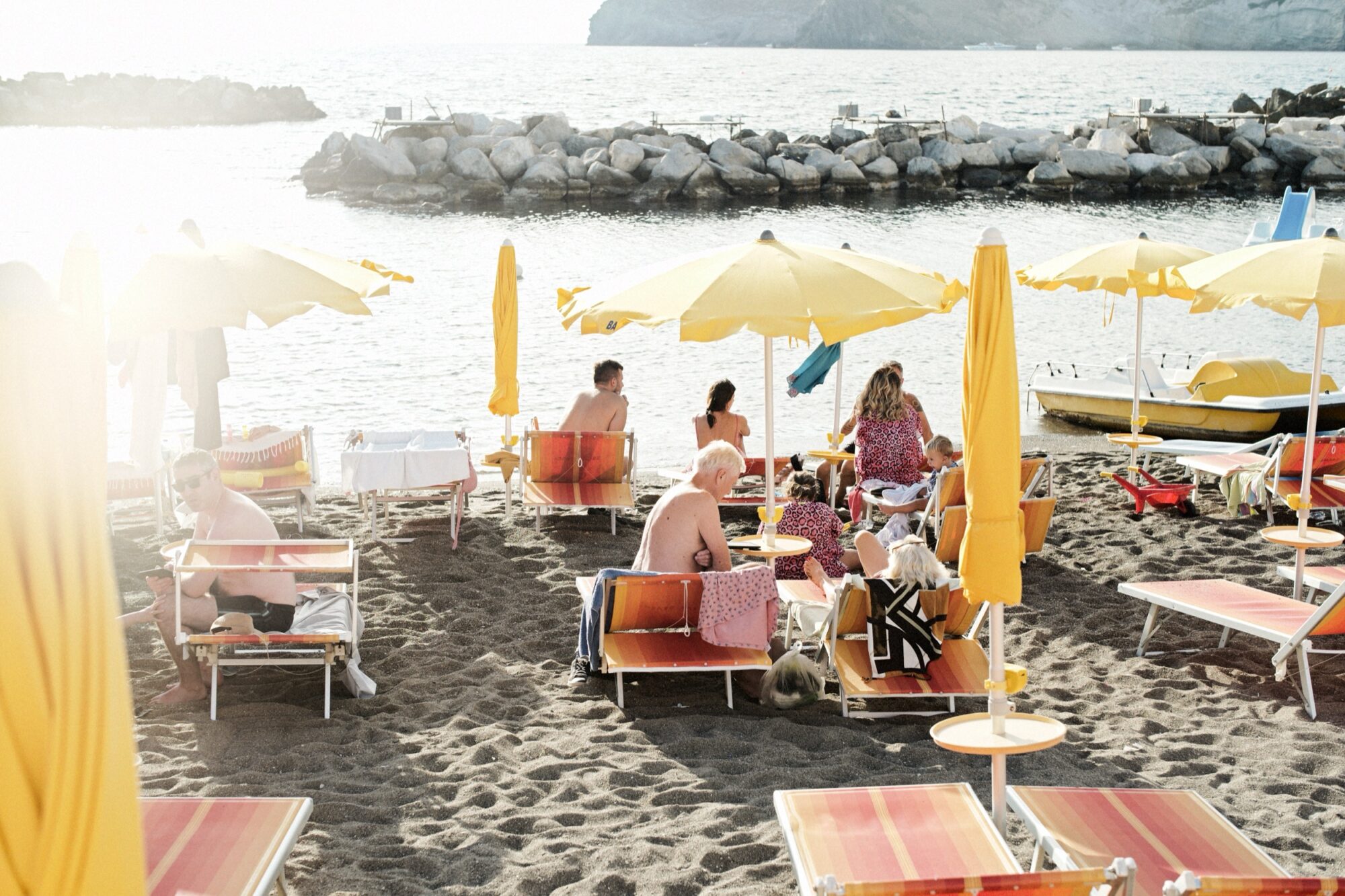 Seattle's best commercial food and hospitality photographer People relax on orange and yellow lounge chairs under yellow umbrellas on a sandy beach by the sea. Some are chatting, others sunbathing, with rocky seawalls and a boat in the water in the background. Sunlight is bright. captured by seattle's best food and lifestyle photographer Brooke Fitts