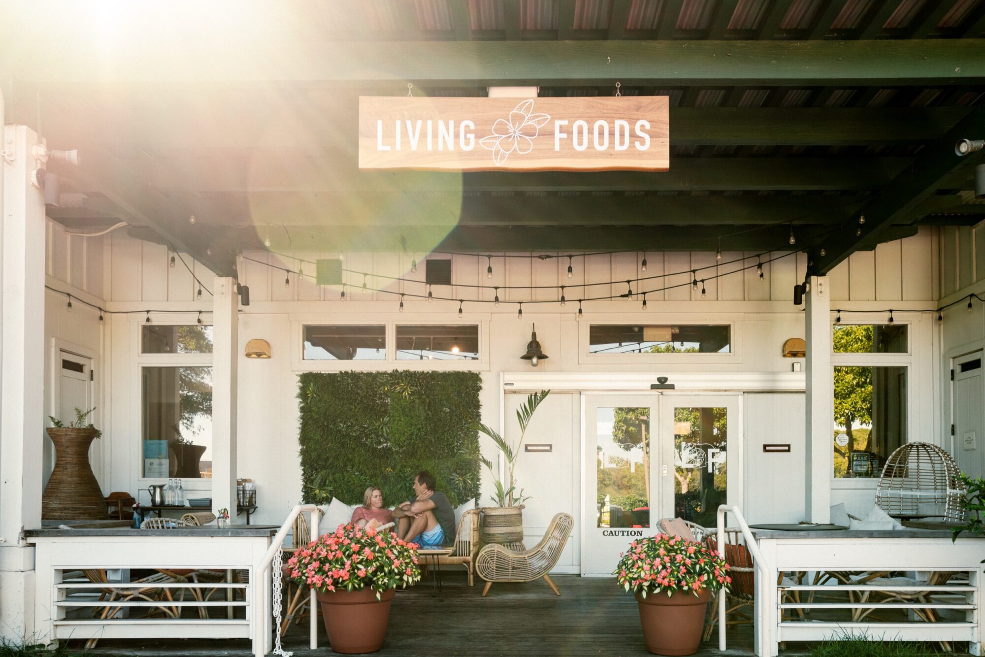 Seattle's best commercial food and hospitality photographer A sunlit porch of a cafe called Living Foods with potted plants, outdoor seating, and string lights. Two people sit at a table, and the entrance features glass doors and greenery. captured by seattle's best food and lifestyle photographer Brooke Fitts