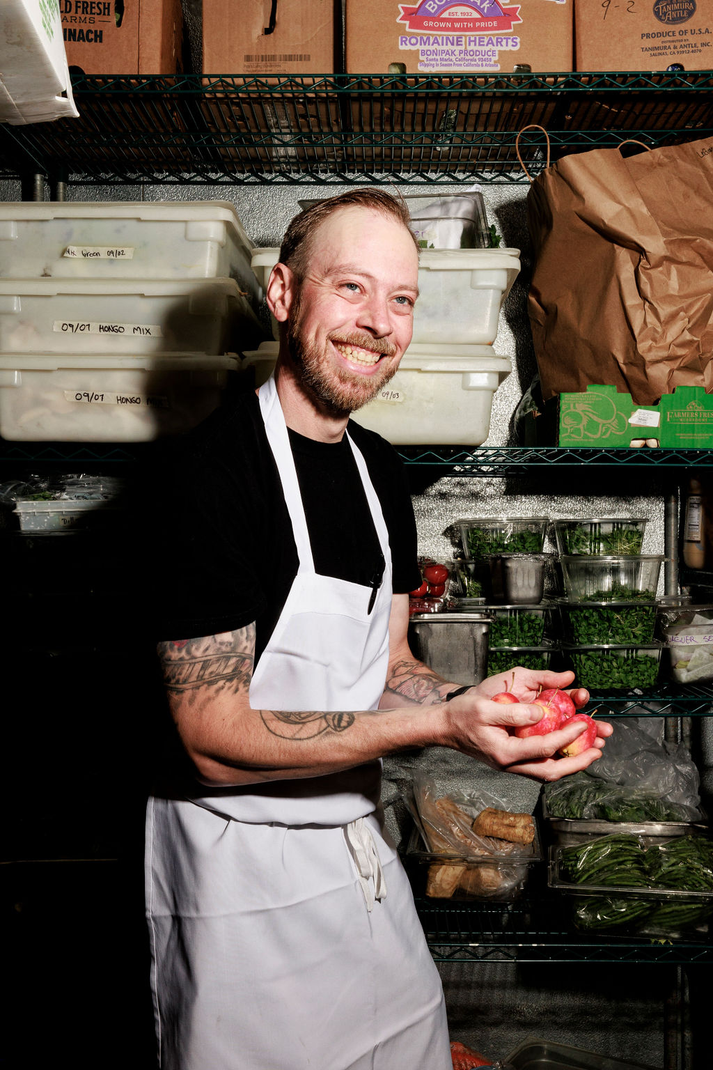 Seattle's best lifestyle potrait photographer A smiling chef wearing a white apron holds a red onion in a commercial kitchen storage area filled with produce, containers, and shelves stocked with fresh ingredients. captured by seattle's best food and hospitality photographer Brooke Fitts