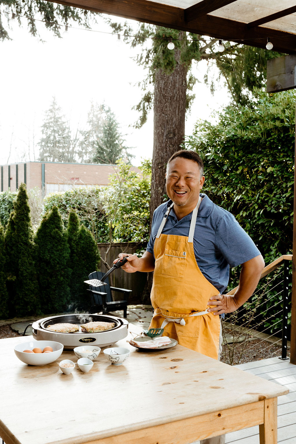Seattle's best lifestyle potrait photographer A man wearing a yellow apron and blue shirt smiles while cooking outdoors at a wooden table with bowls and cooking utensils, surrounded by greenery and trees. captured by seattle's best food and hospitality photographer Brooke Fitts